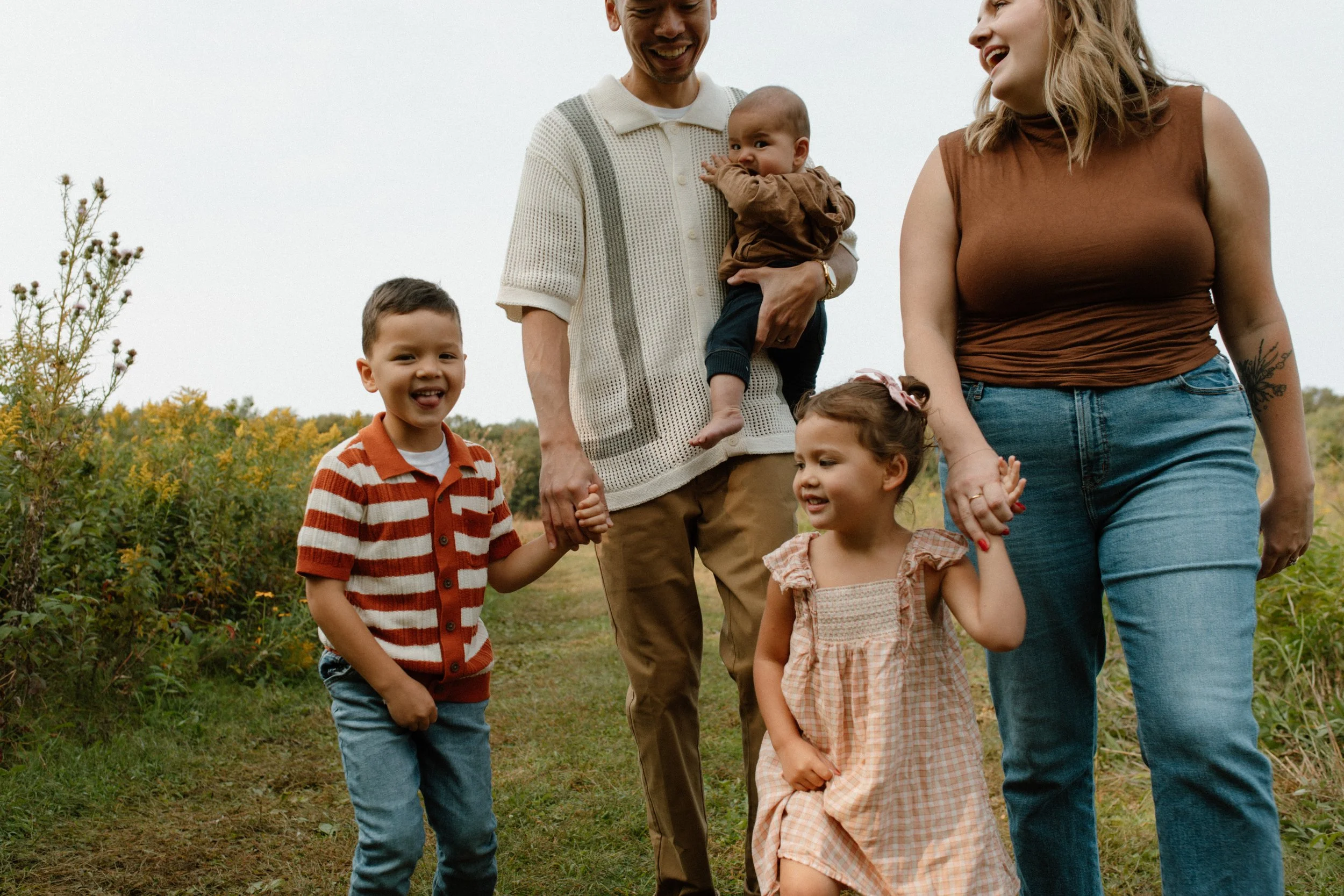 A diverse family of six, including two adults and four children, holding hands and walking outdoors in a green field with yellow flowers, enjoying a sunny day.