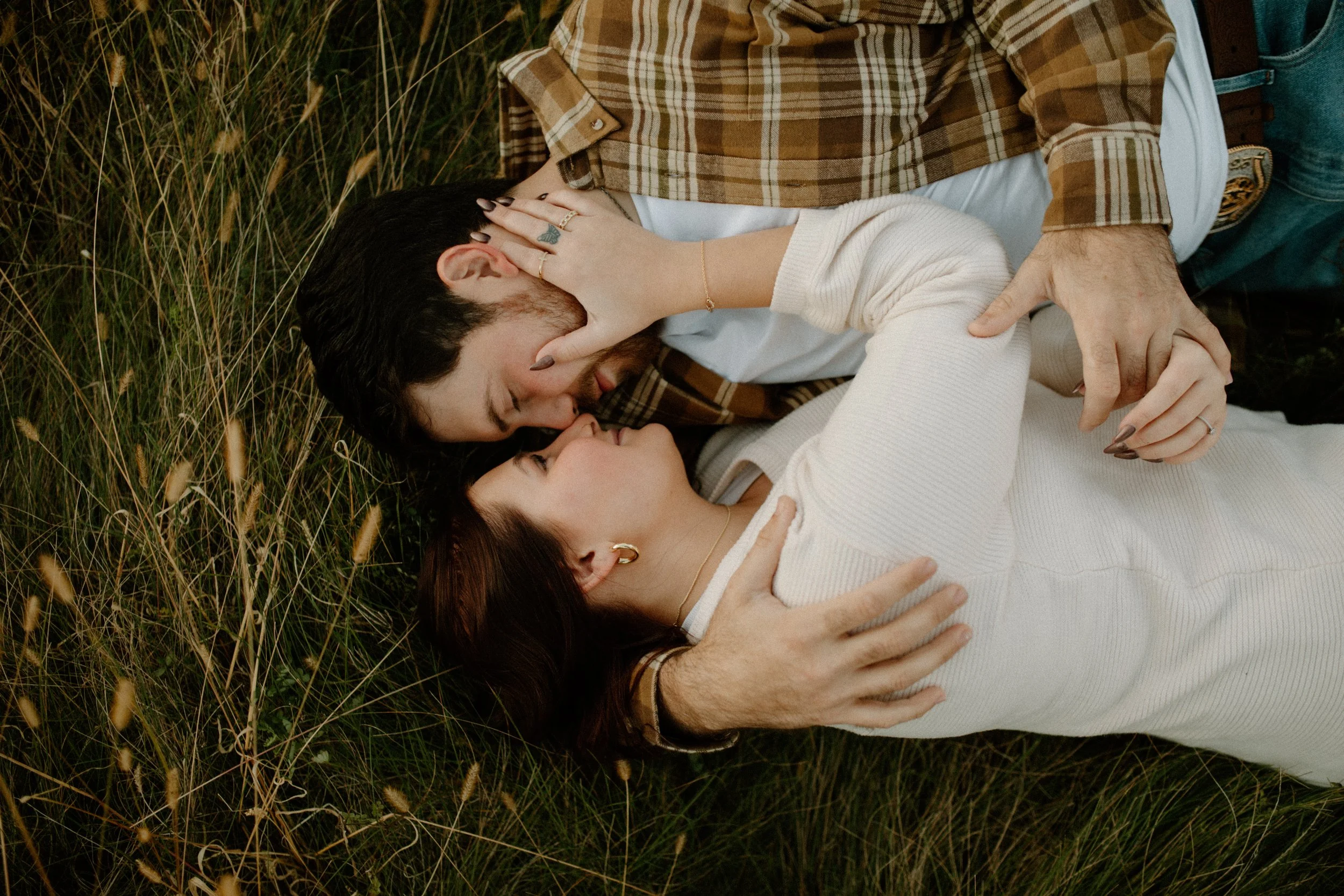Two women lying on grass, one with dark hair and the other with red hair, sharing an intimate moment, with the woman with dark hair touching the neck of the woman with red hair.
