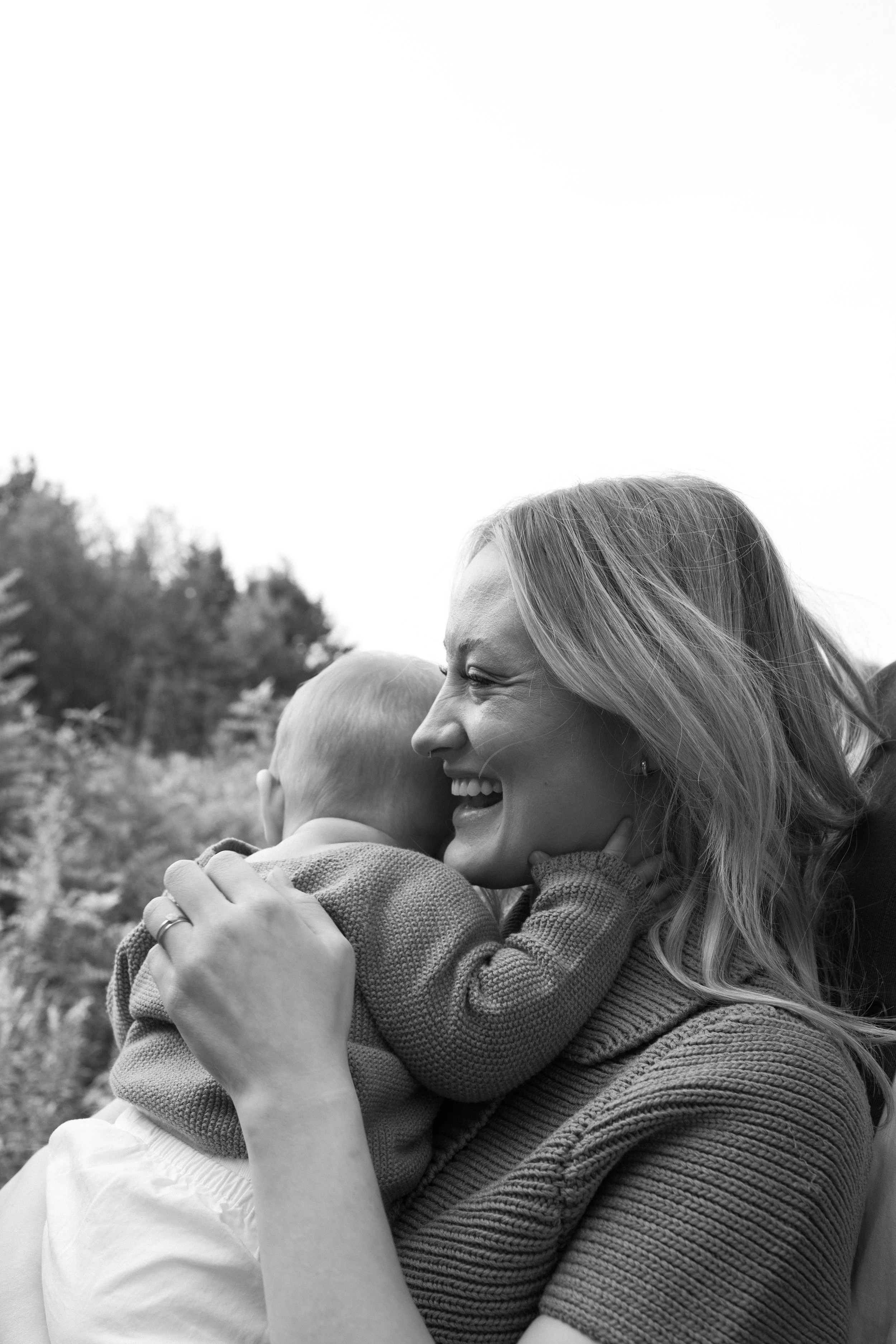 A woman with long hair and a young child are embracing and smiling outdoors with trees in the background.