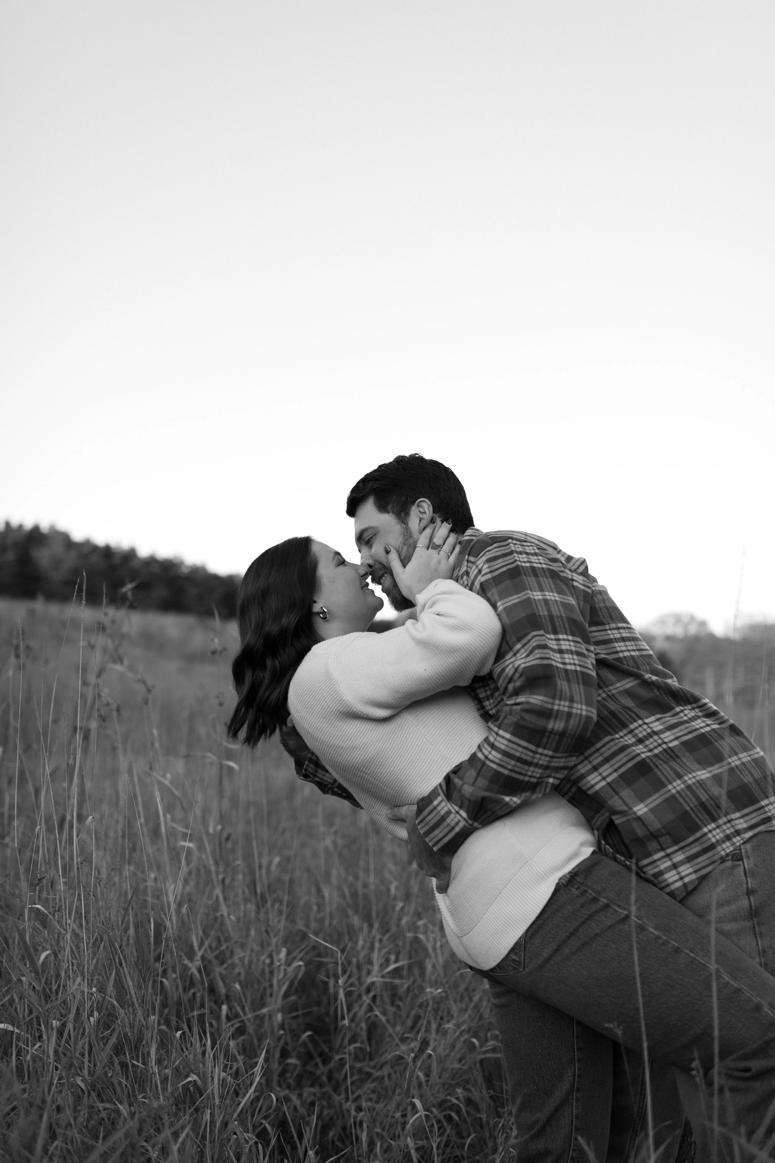 A black-and-white photo of a couple in an outdoor grass field, with the man dipping the woman backwards and both smiling and gazing into each other's eyes.