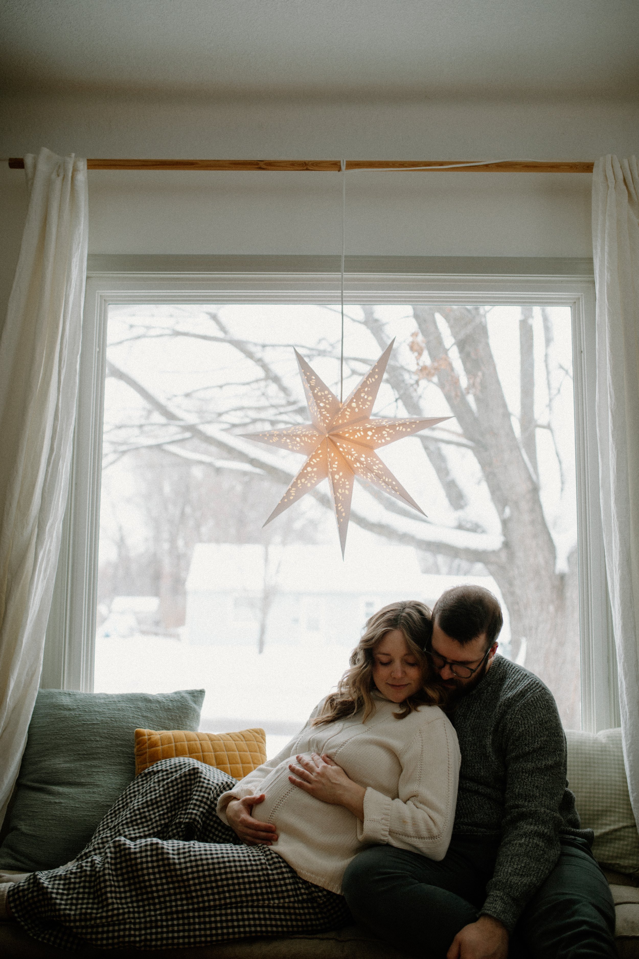 A couple sitting on a sofa by a window, with the woman pregnant and the man holding her belly, in a cozy, warmly-lit room with a hanging star-shaped lantern and cushions.