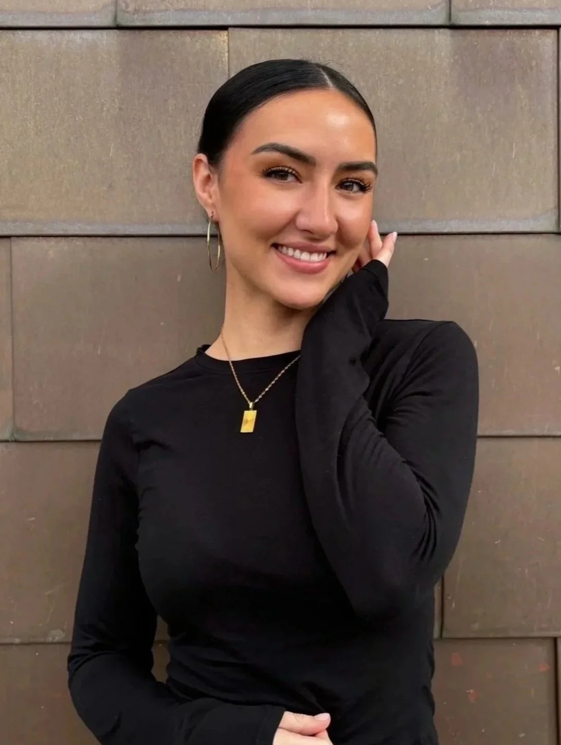 Young woman smiling, wearing a sleeveless white top and standing against a metallic wall background.