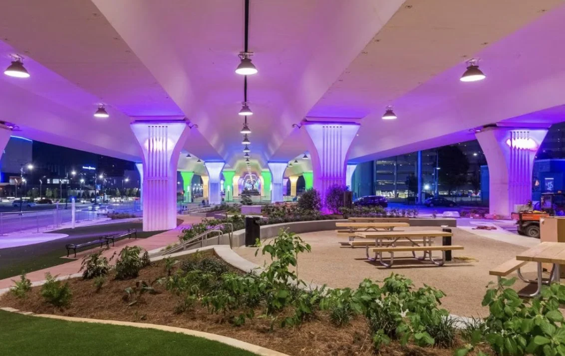 Underpass with columns illuminated in purple and green lights, benches, picnic tables, and landscaped plants beneath, at night.