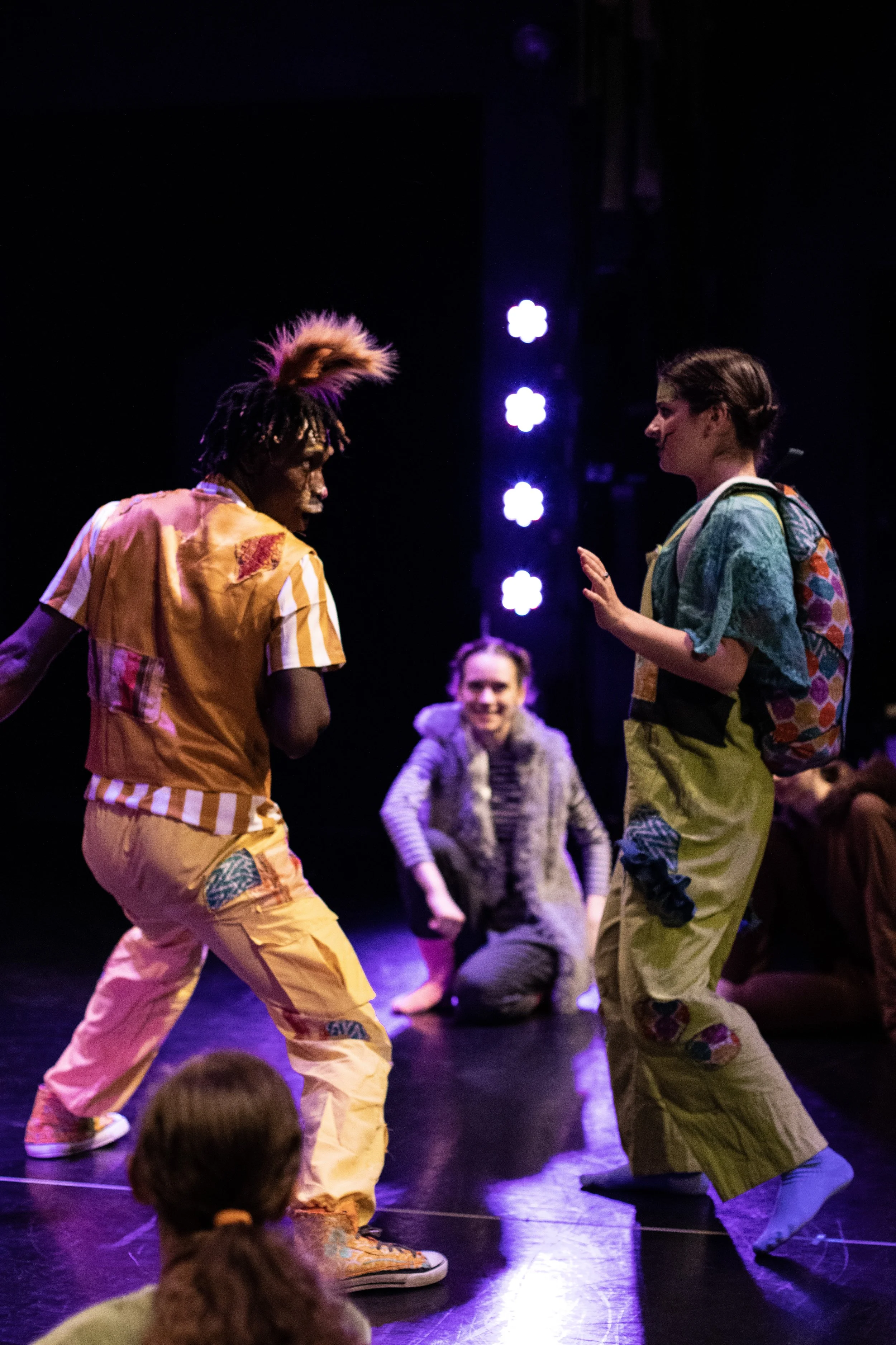 Two actors perform on stage, one wearing rabbit ears and a colorful patchwork outfit, and the other green overalls and a multicolored backpack that looks like a tortoise shell.