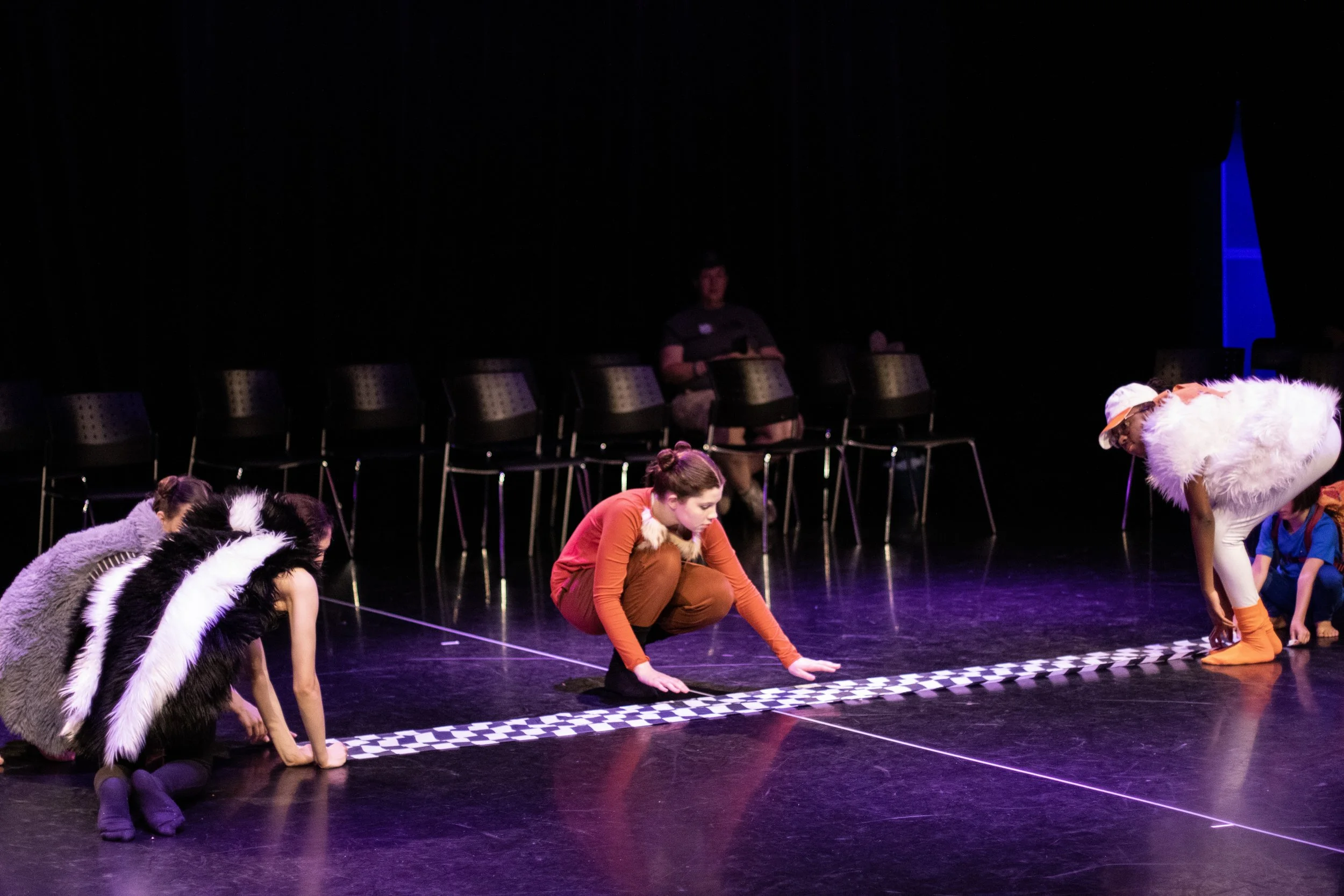 Children in animal costumes preparing for a performance on a dark stage with chairs in the background.