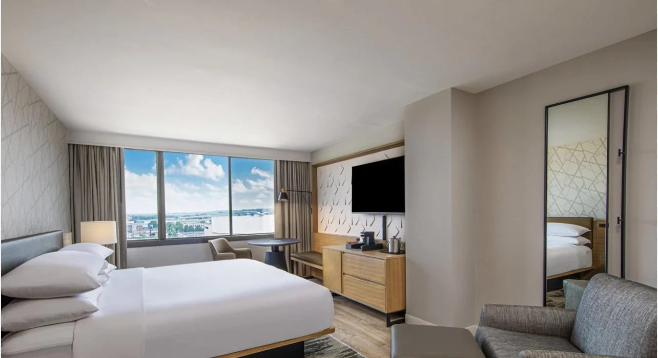 A modern hotel room with a large bed, a living area with a chair and sofa, a flat-screen TV, a window with a city view, and a sliding mirror door to a bathroom.