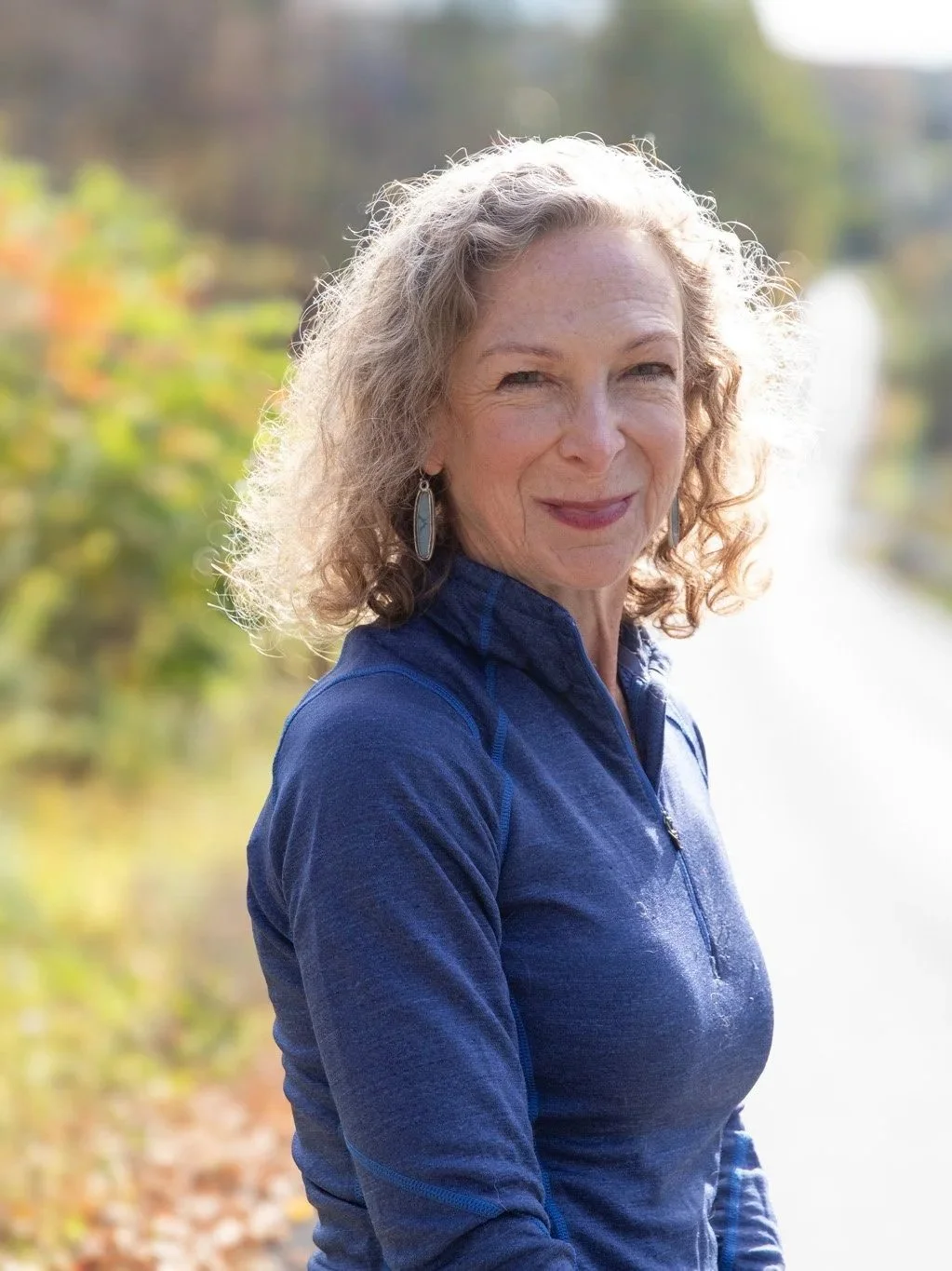 A woman with curly gray hair wearing a blue jacket outdoors, smiling at the camera with a background of green and yellow foliage and a body of water.