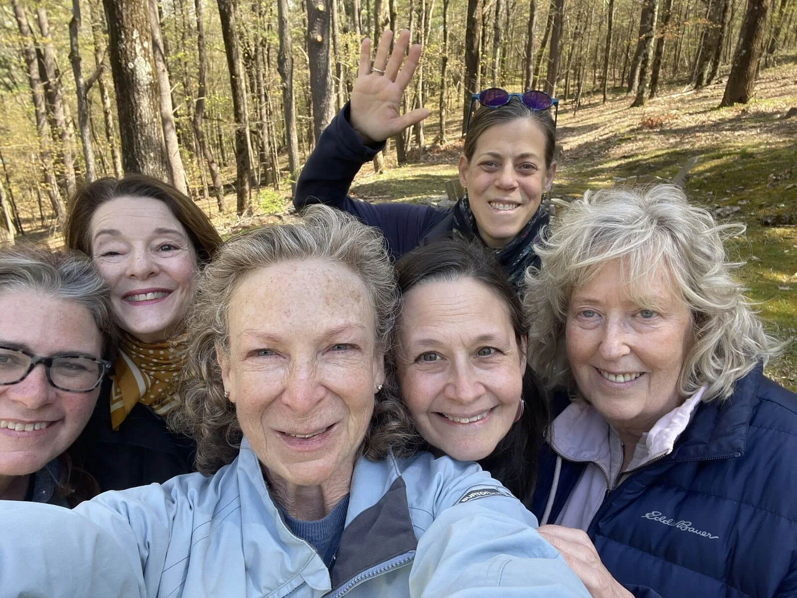 Six women taking a group selfie in a wooded area during autumn, smiling and posing happily.
