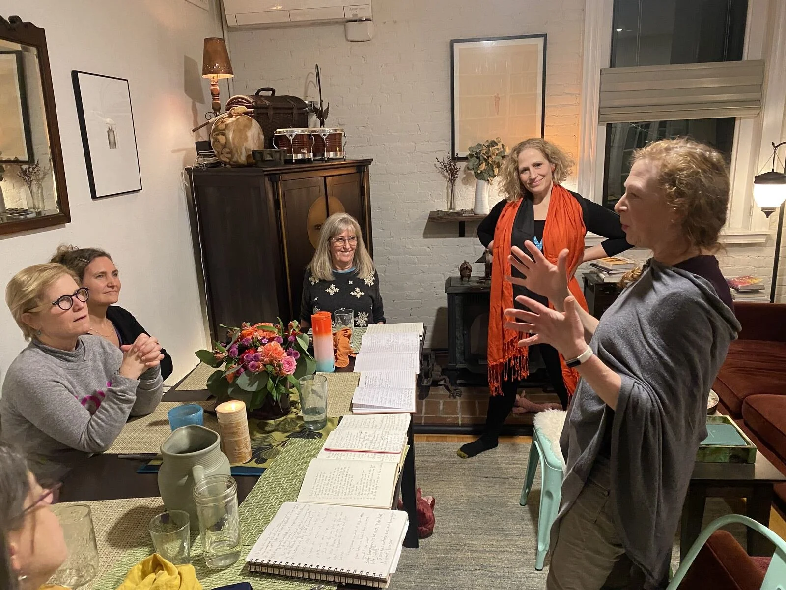 Group of women gathered around a dining table with notebooks, glasses, and flowers, engaged in conversation in a cozy, well-lit living room.