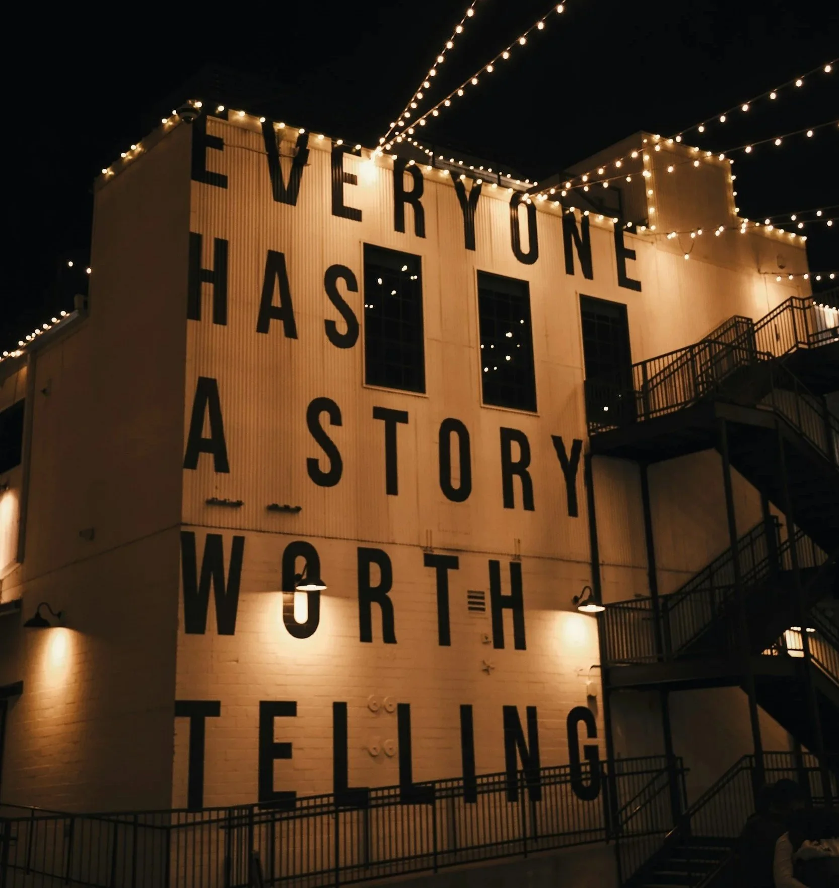Nighttime view of a building with large black lettering spelling out 'EVERYONE HAS A STORY WORTH TELLING,' decorated with string lights along the roofline and a fire escape on the right side.