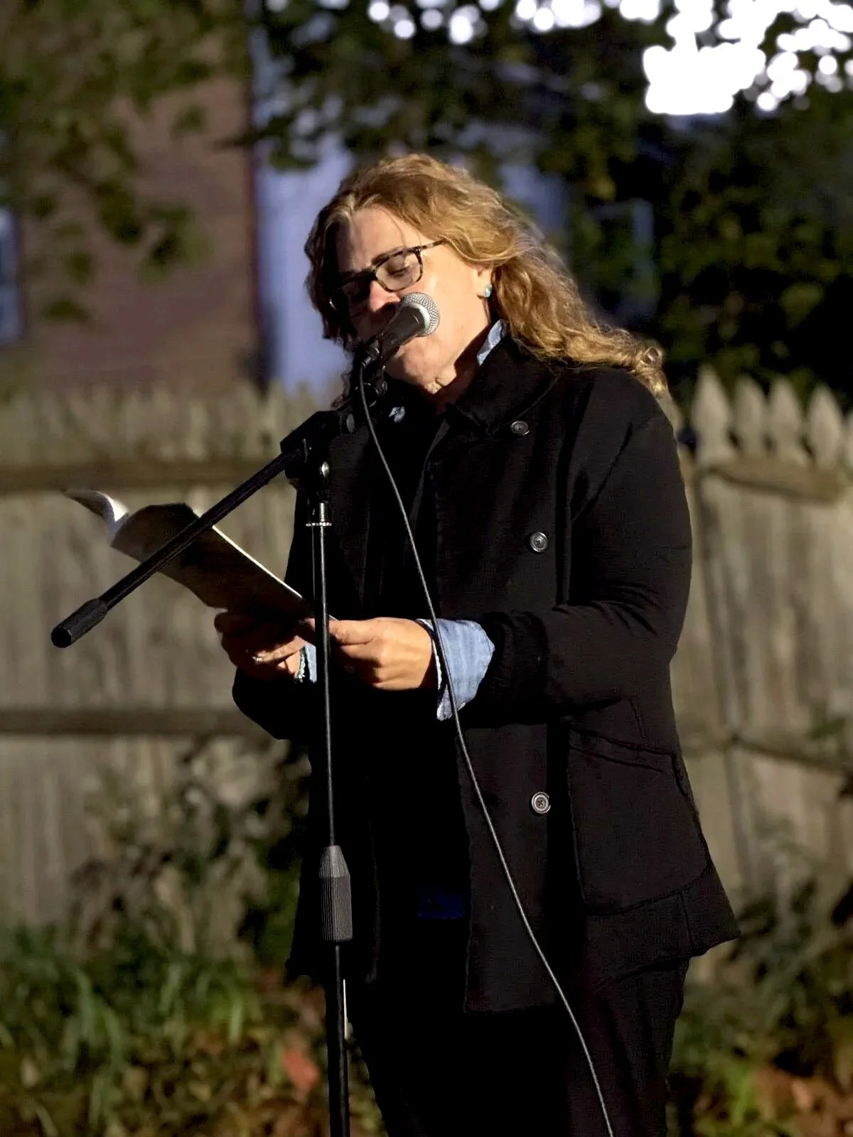 Woman with glasses and curly hair reading from a tablet at a microphone outdoors during evening.