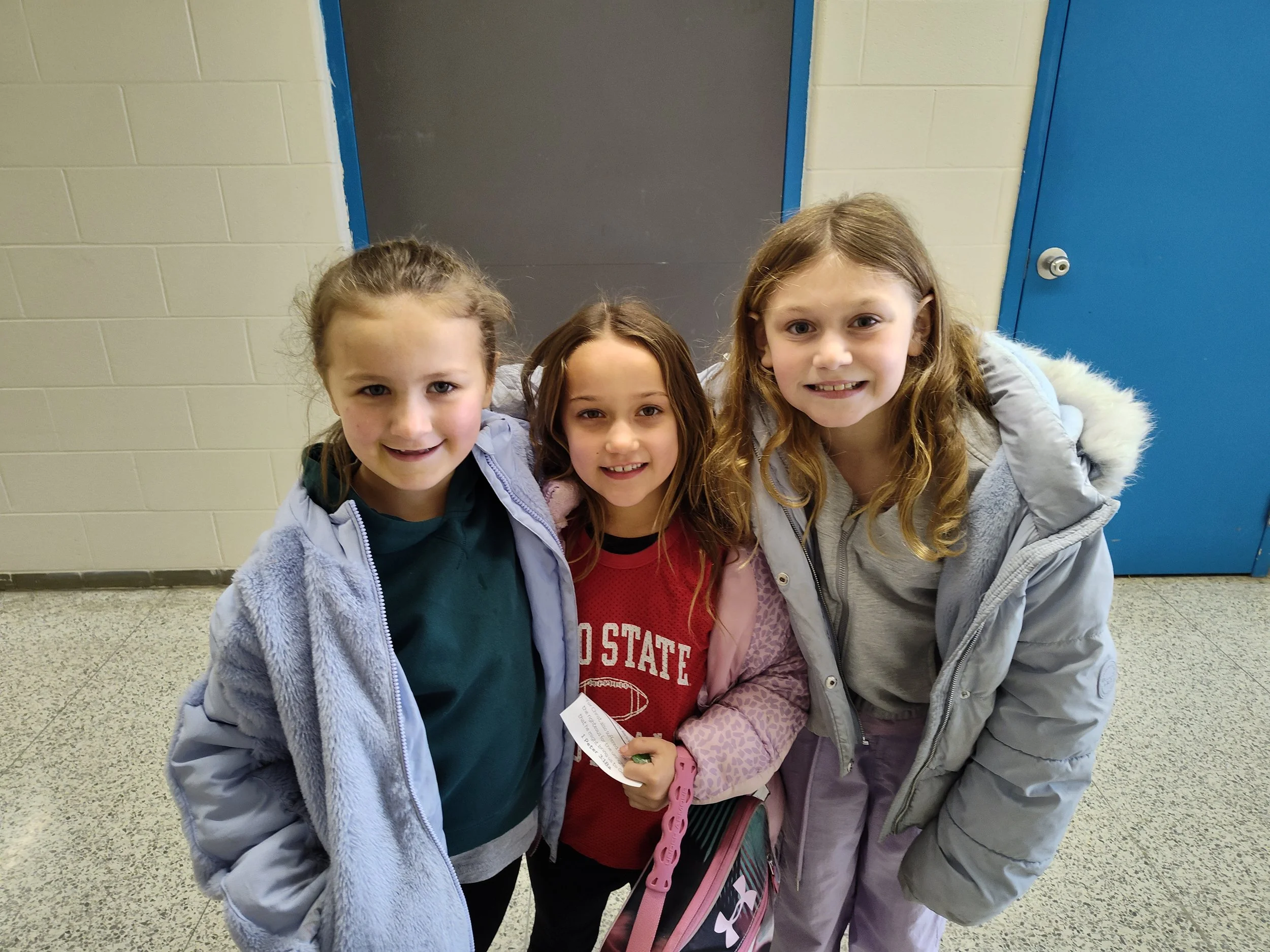 A classroom with children sitting at a long table, some raising their hands, and an adult woman sitting at the back.