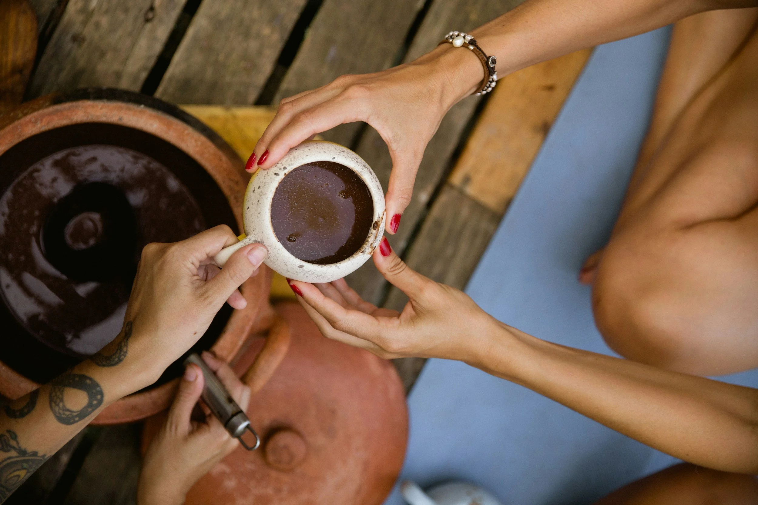 Two hands holding a white mug filled with coffee, one hand with red nail polish and a bracelet, the other with tattoos. A portion of a tattooed arm is visible. The background includes a wooden surface and a blue wall.