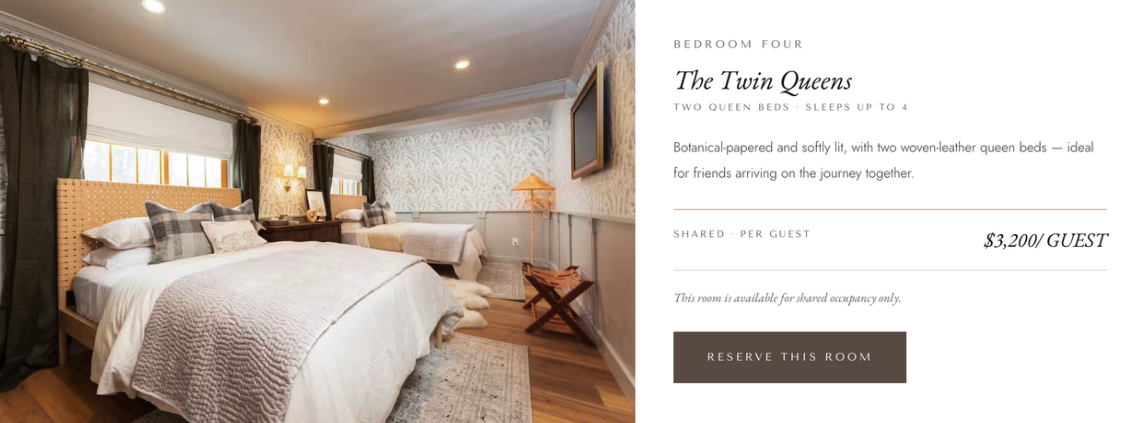 Photo of a bedroom with two queen beds, featuring a wooden headboard, multiple pillows, and a cozy blanket. The room has windows with dark curtains, wooden flooring, and a wall with patterned wallpaper. There is a small wooden side table, a lamp, and a painting on the wall.