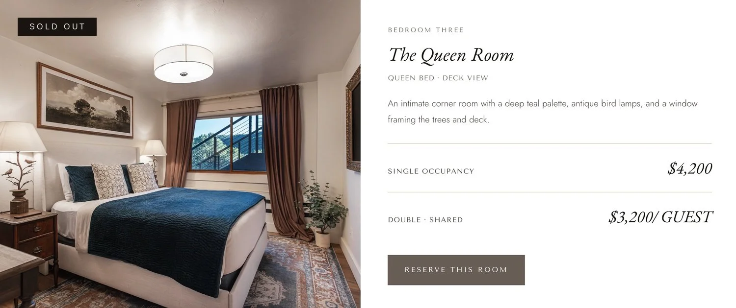 A cozy bedroom with a queen-sized bed dressed in navy and white bedding, framed by mocha-colored curtains and a window with a deck railing visible outside. The room features side tables with table lamps and wall art above the bed.