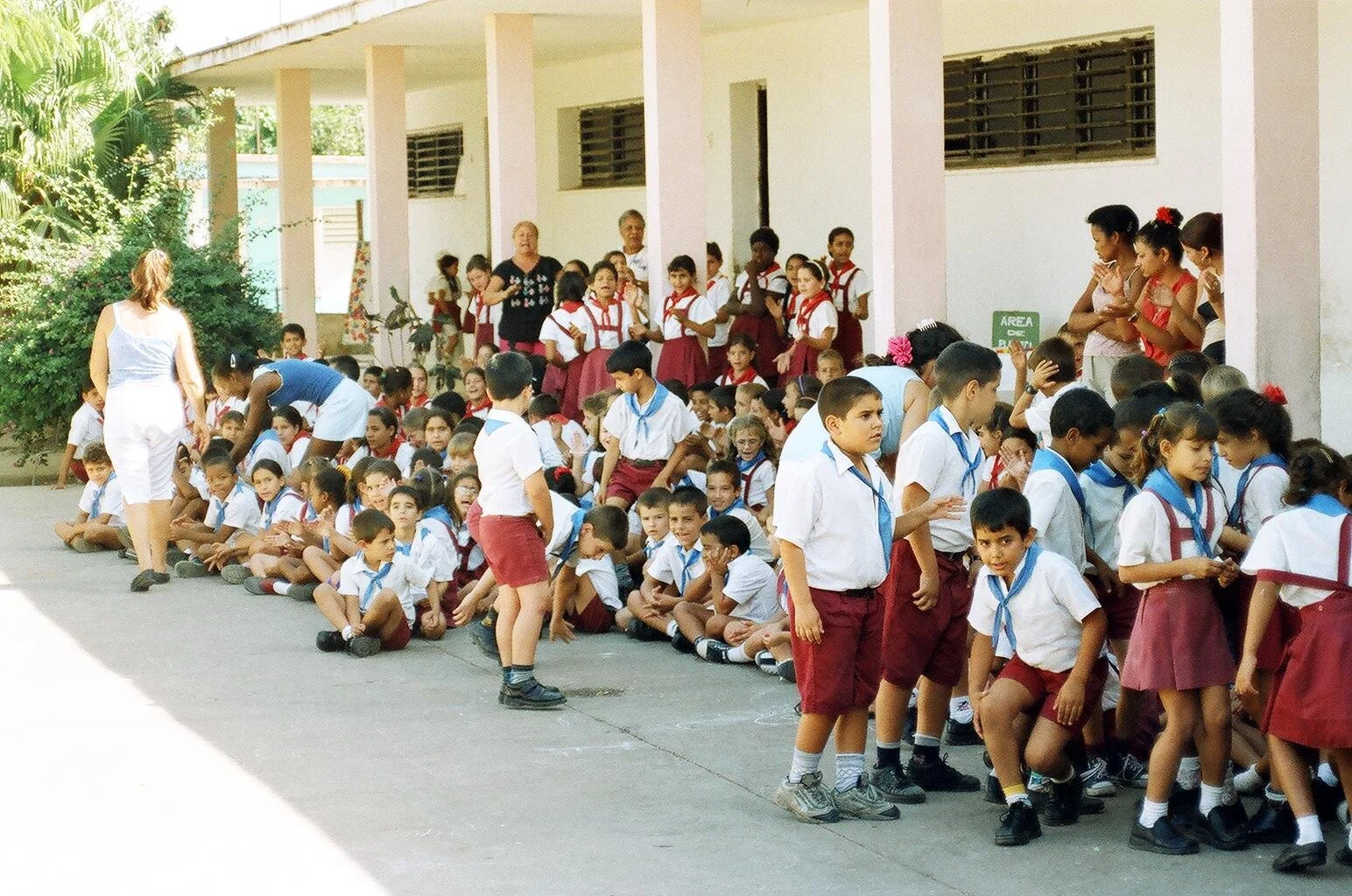 Group of elementary school children in uniform sitting and standing outside a school building, with teachers supervising.