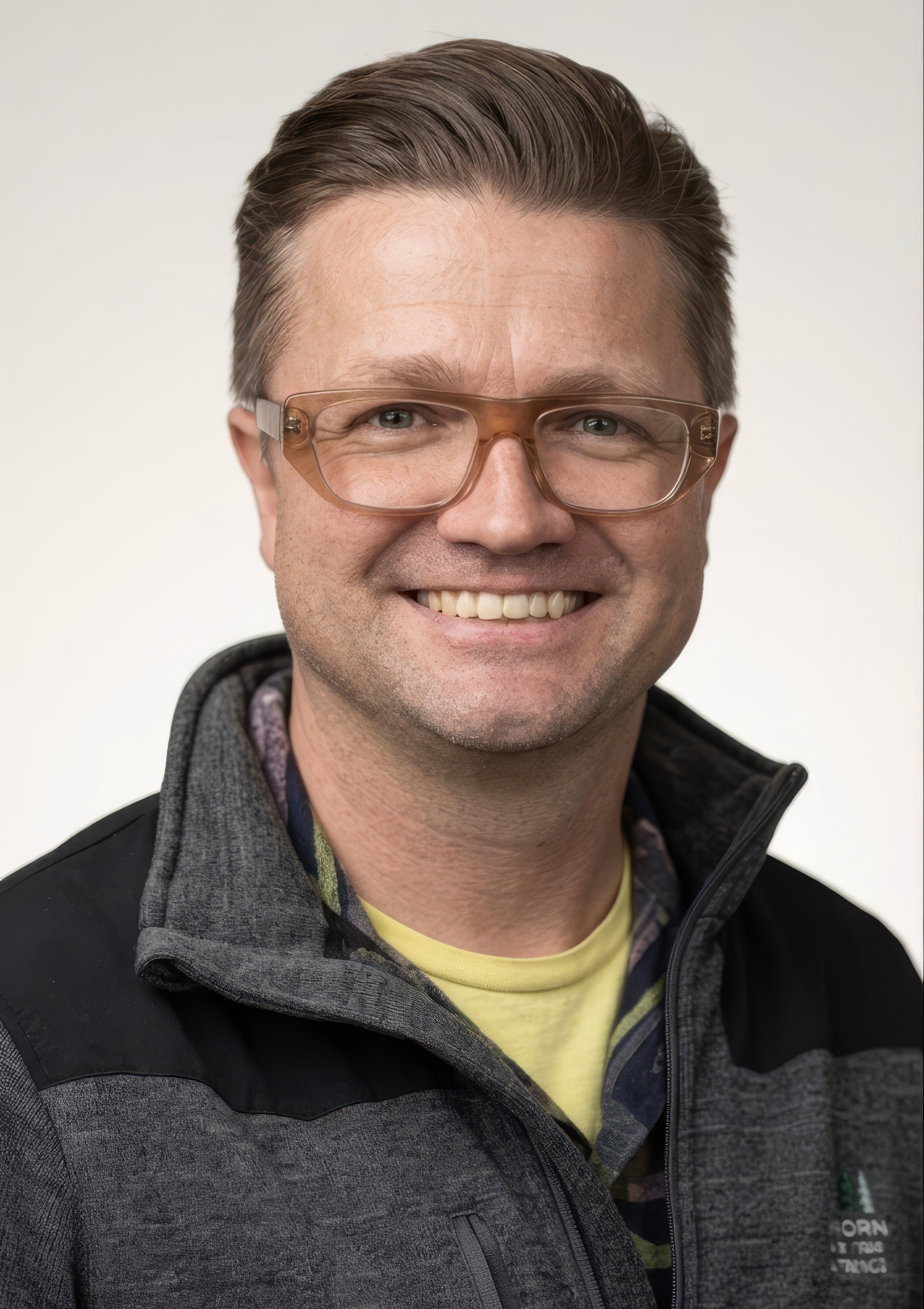 A man with glasses smiling, wearing a black and grey jacket over a yellow shirt, with short brown hair.