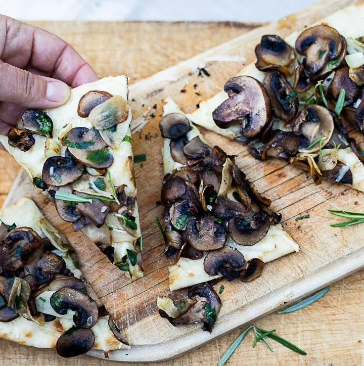 A hand is taking a slice of mushroom pizza topped with sliced mushrooms and herbs from a wooden cutting board.