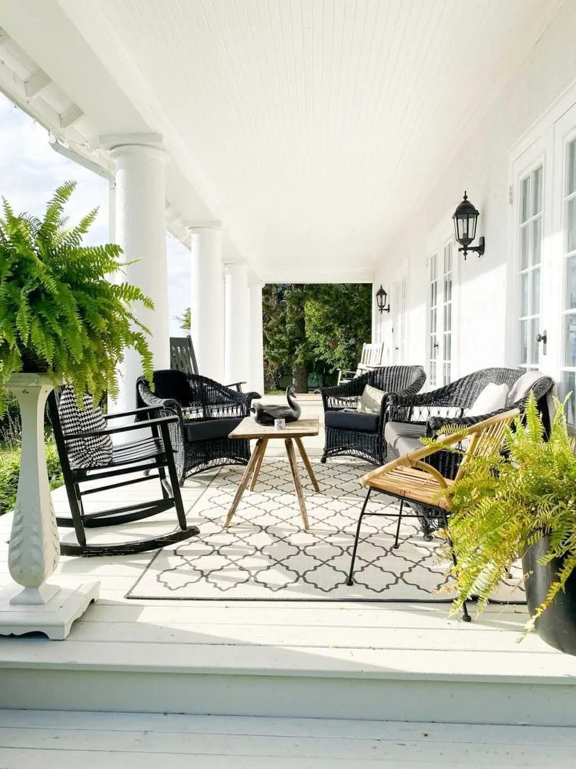 A spacious outdoor porch with white walls and ceiling, furnished with black and tan wicker chairs, a wooden coffee table, and a patterned rug. Green plants are on either side, and black lantern-style wall lights are mounted on the wall next to multip