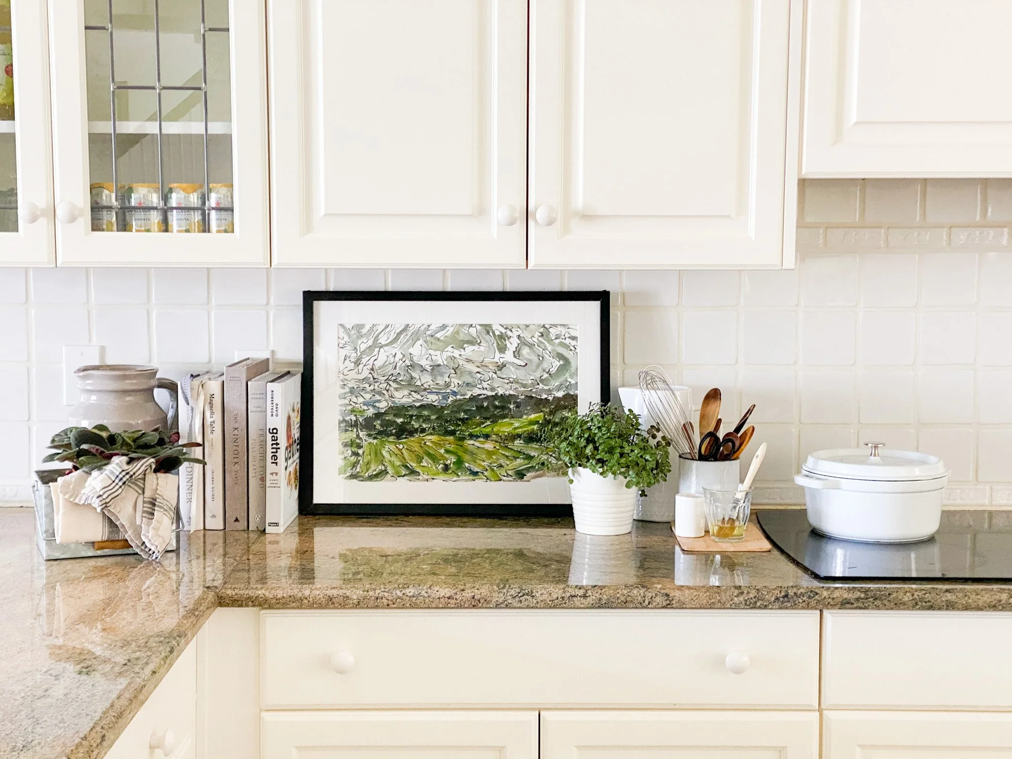Deconstructed kitchen countertop with books, framed artwork, potted plant, utensils, oil, container, and stovetop.