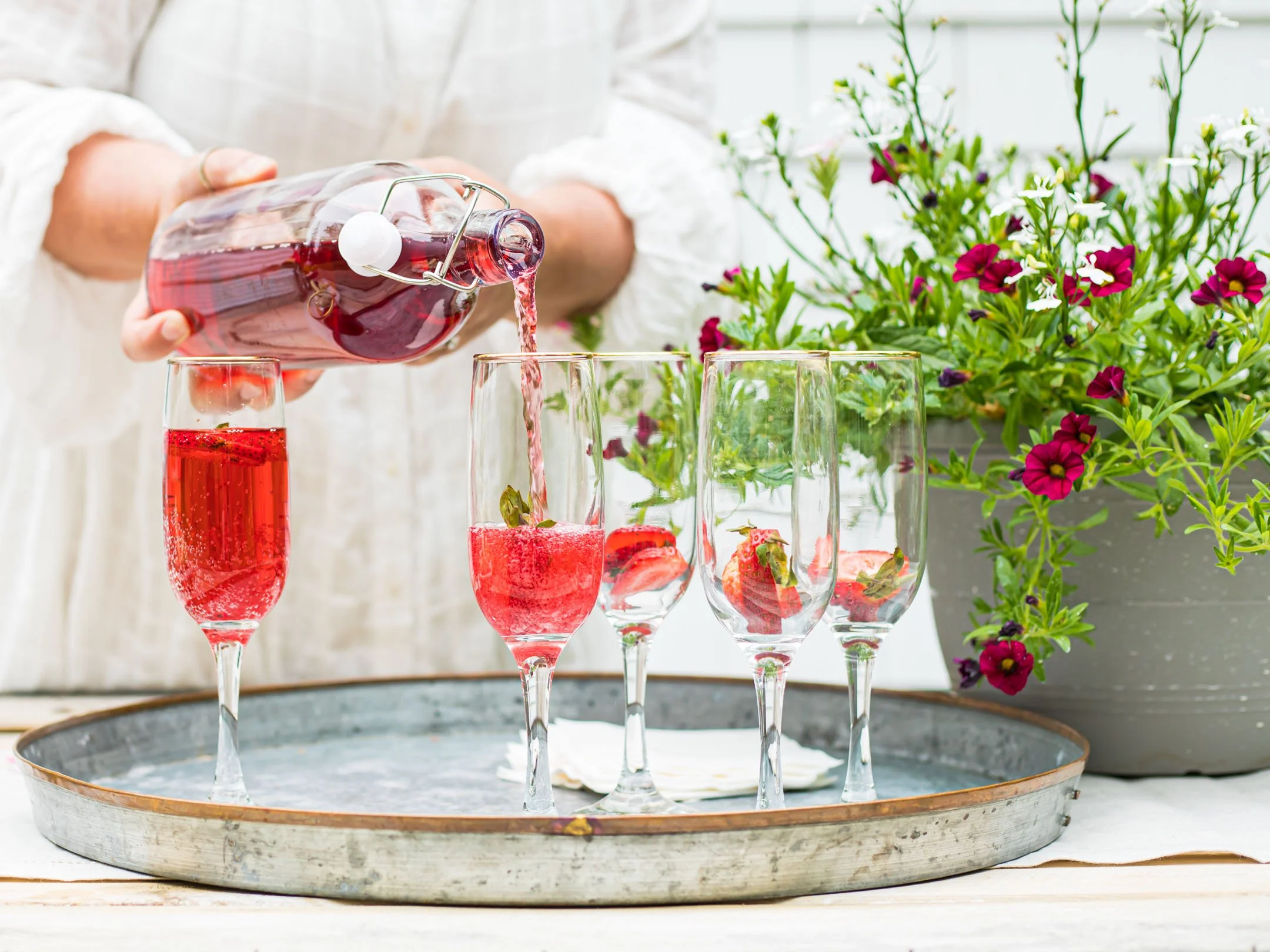 Person pouring pink sparkling wine into champagne flutes with strawberries, on a tray, next to a pink and white flowering plant.