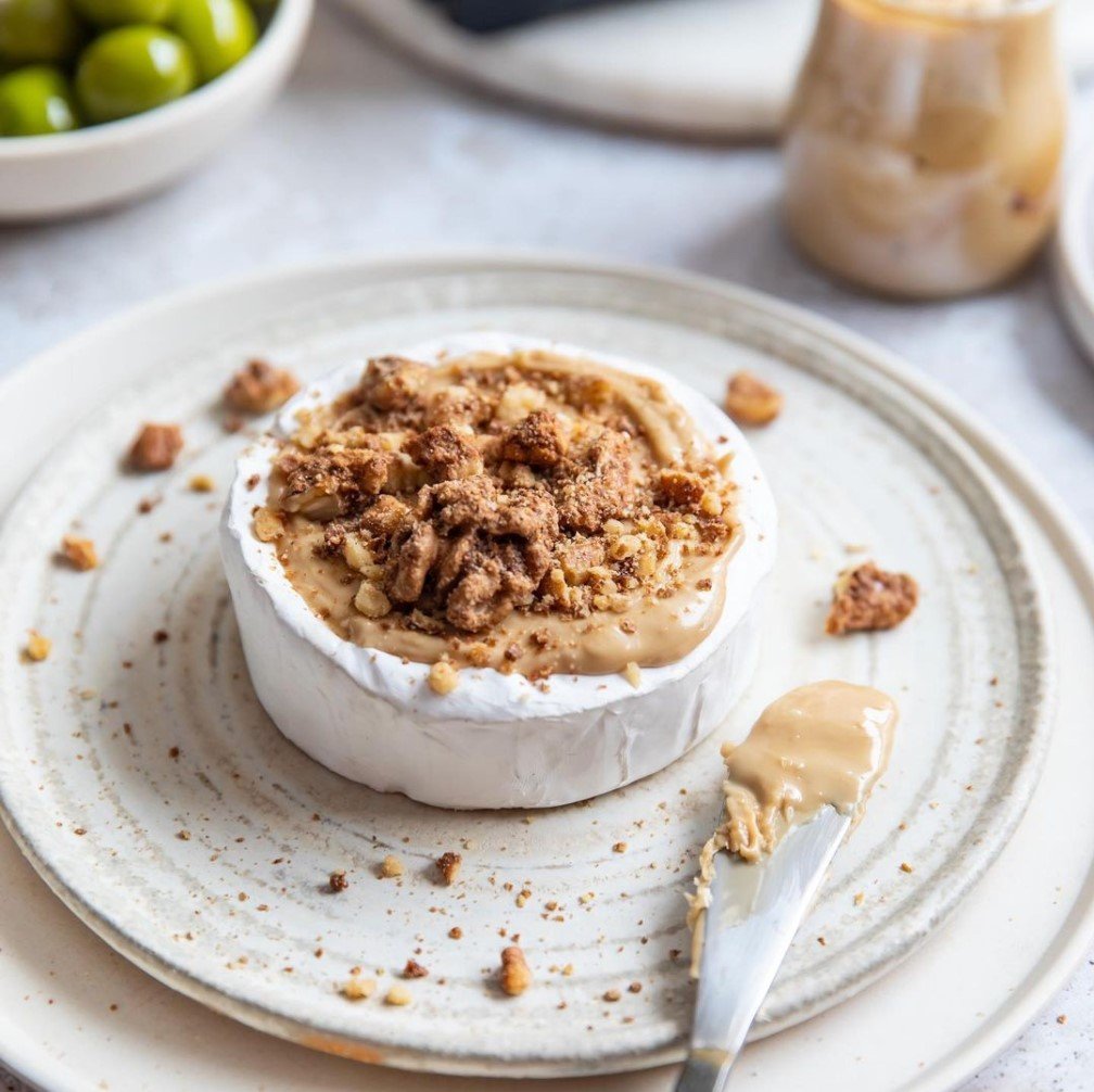 A baked brie cheese round topped with crushed cookies or crackers on a plate with a spoon, surrounded by various ingredients and dishes.