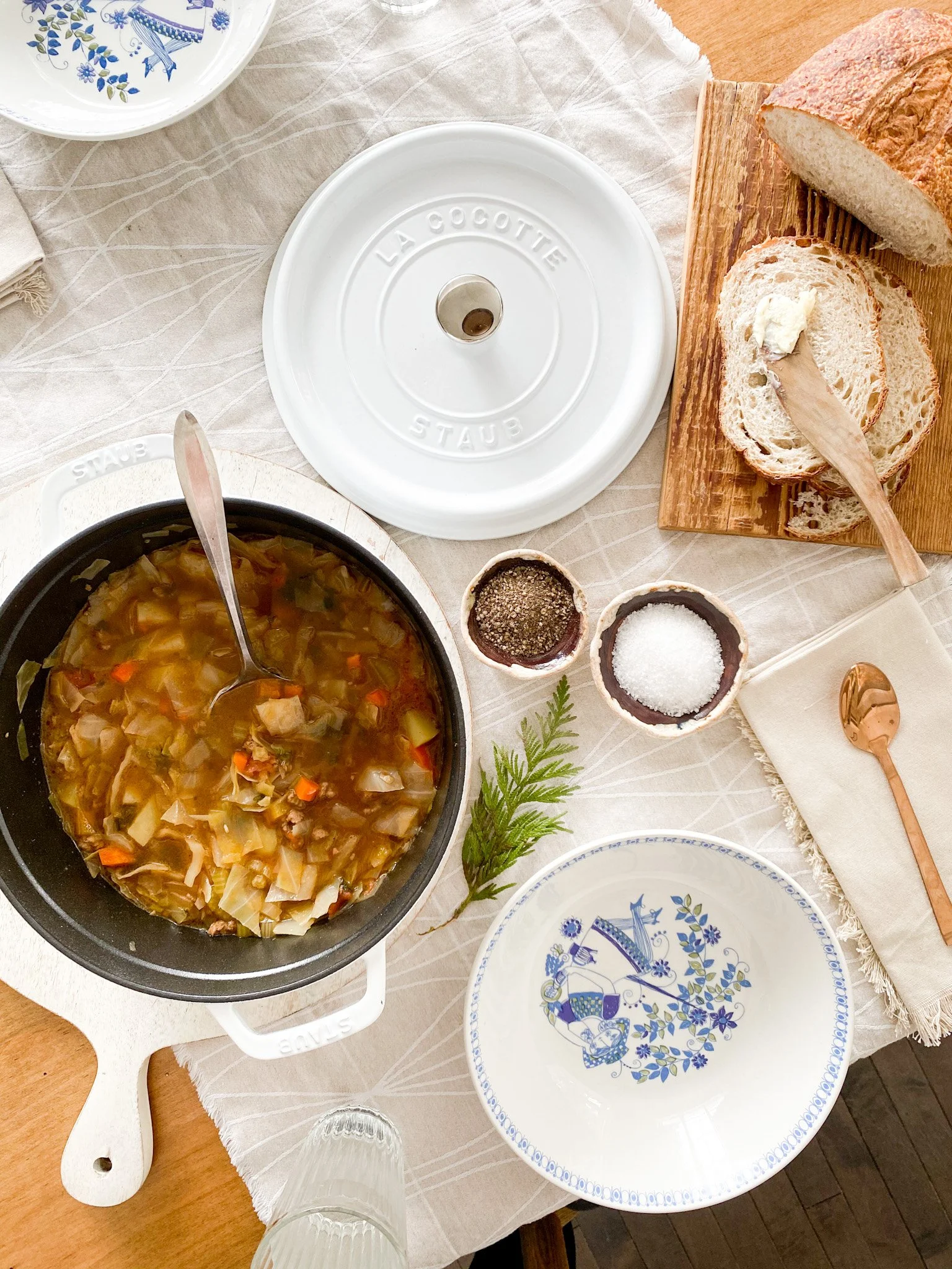 A table with a pot of vegetable soup, slices of bread with butter, salt and pepper shakers, and decorative plates with blue floral and figure designs.