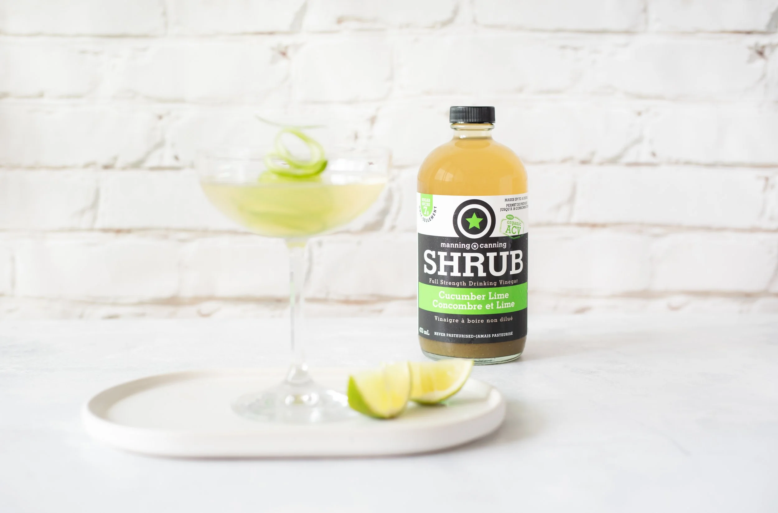 A bottle of Manning Canning Shrub Cucumber Lime drinking vinegar on a white surface with lime wedges and a cocktail glass containing lime peel, against a white brick wall background.