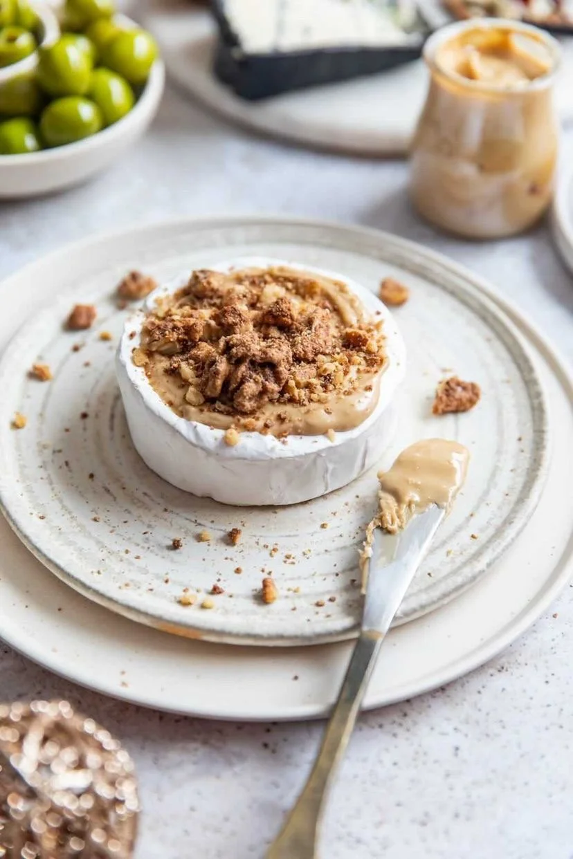 A wheel of brie cheese topped with caramel and crushed cookies on a white plate, with a butter knife spread with caramel in front, and a jar of caramel sauce in the background.