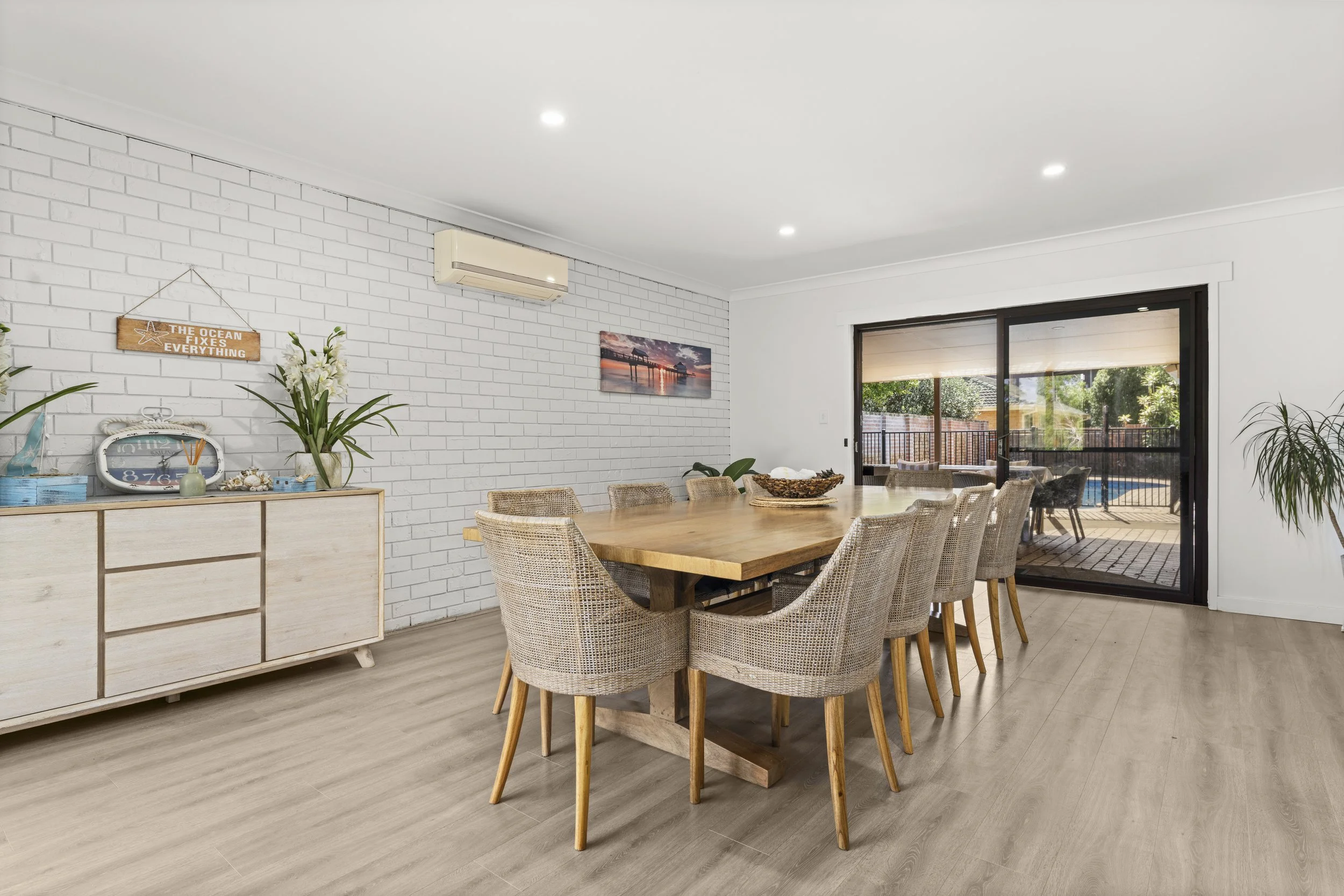 A dining room with a large wooden table and ten woven chairs, a sideboard with decor, and sliding glass doors leading to an outdoor patio with a pool.