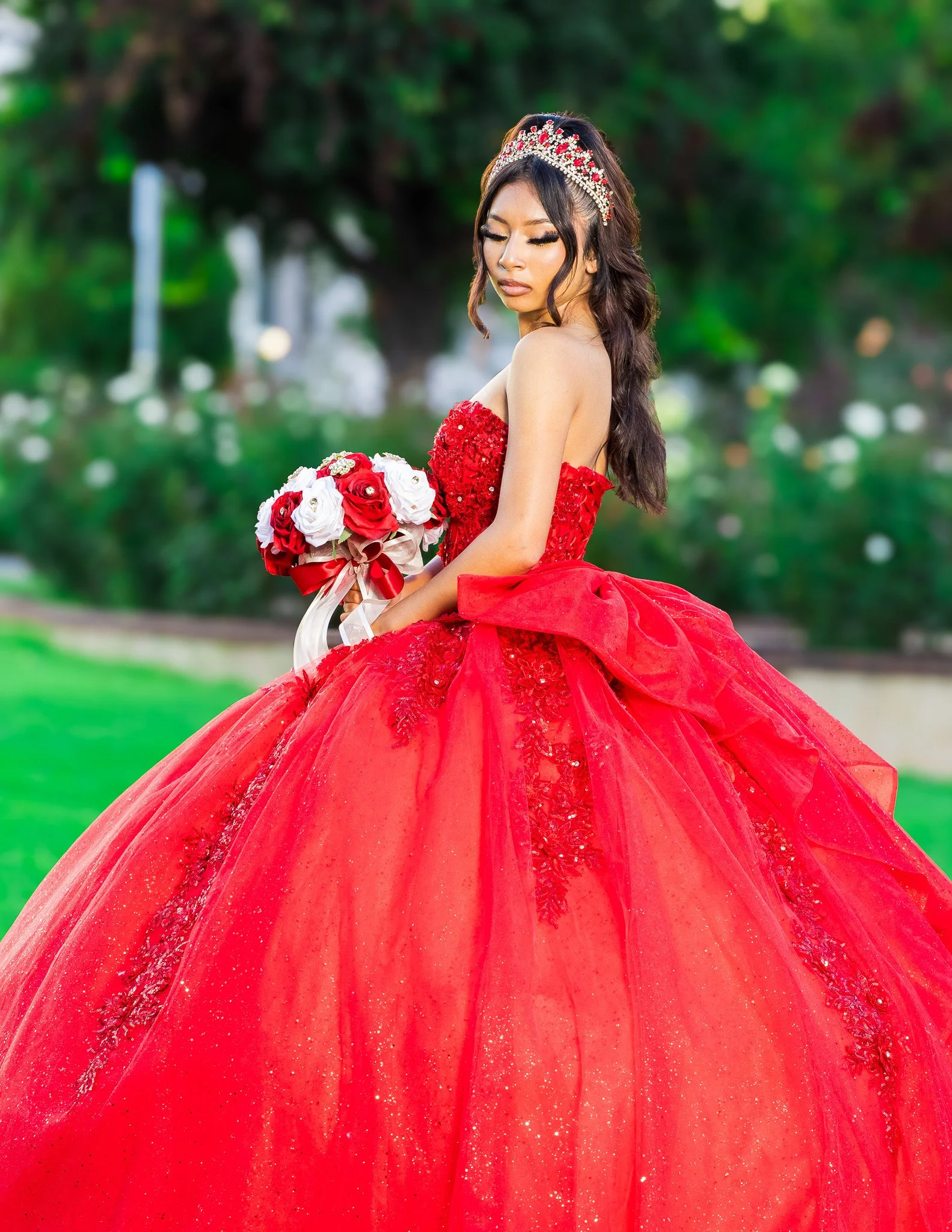 Woman in a sparkling red ball gown and jeweled tiara holding a red and white rose bouquet outdoors