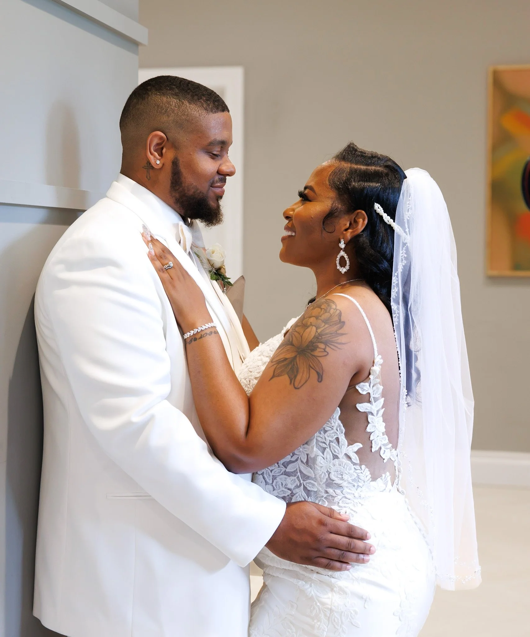 A bride and groom standing close together indoors, touching foreheads and looking into each other's eyes. The groom is dressed in a white suit with a boutonniere, while the bride wears a lace wedding dress and veil, with tattoos on her arm and large dangling earrings.