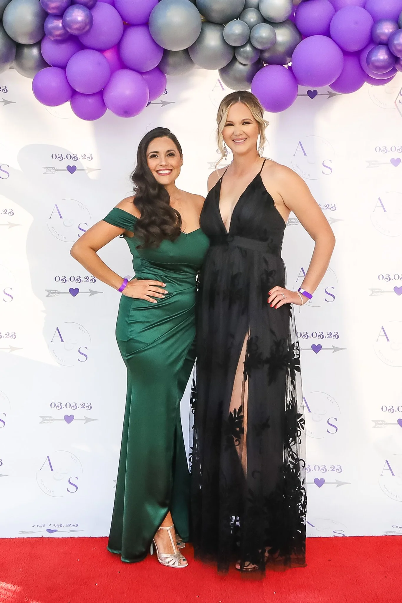 Two women in evening dresses standing on a red carpet at a formal event with purple balloons and a white backdrop with purple text and date, smiling for the camera.