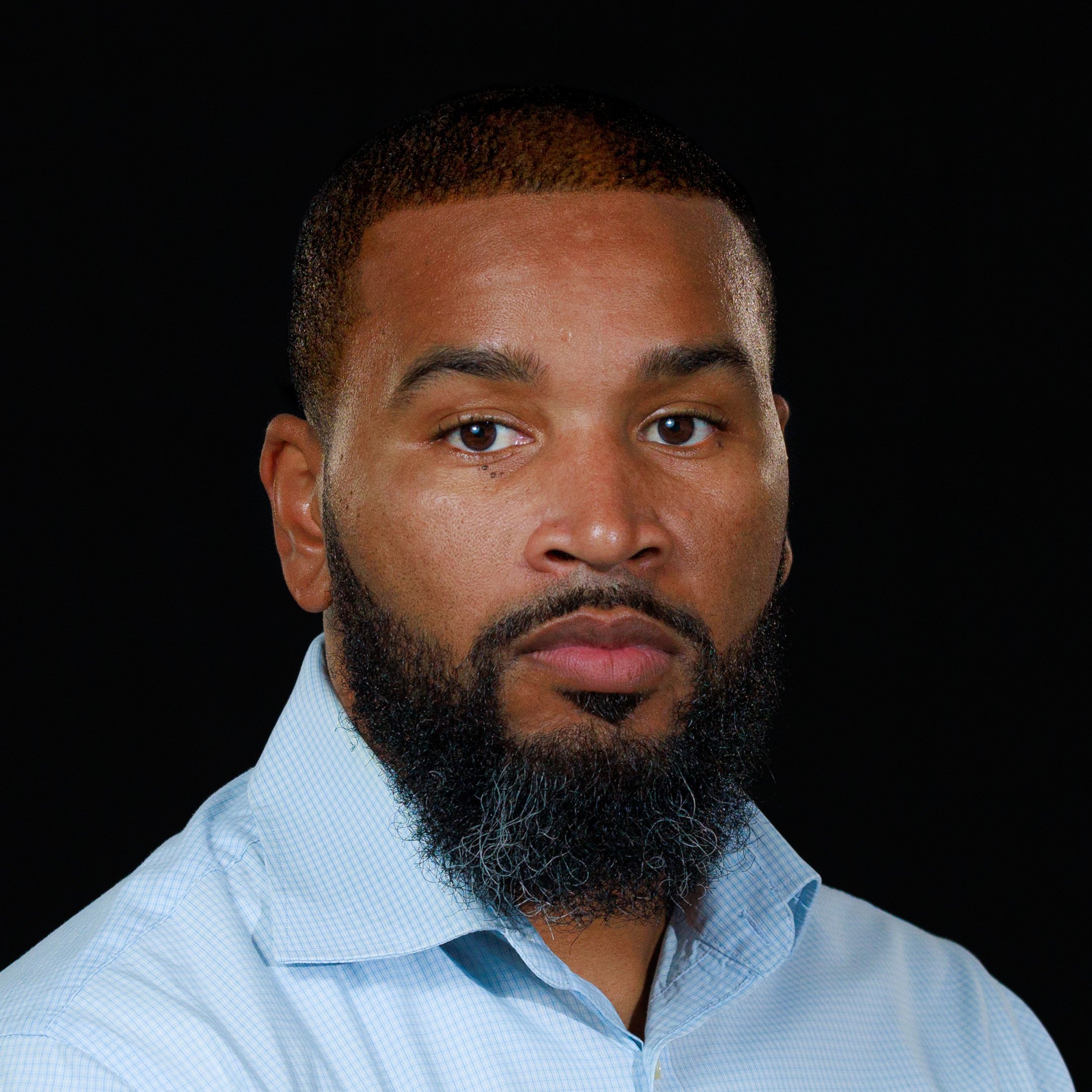 Close-up portrait of a bearded Black man wearing a light blue shirt against a black background