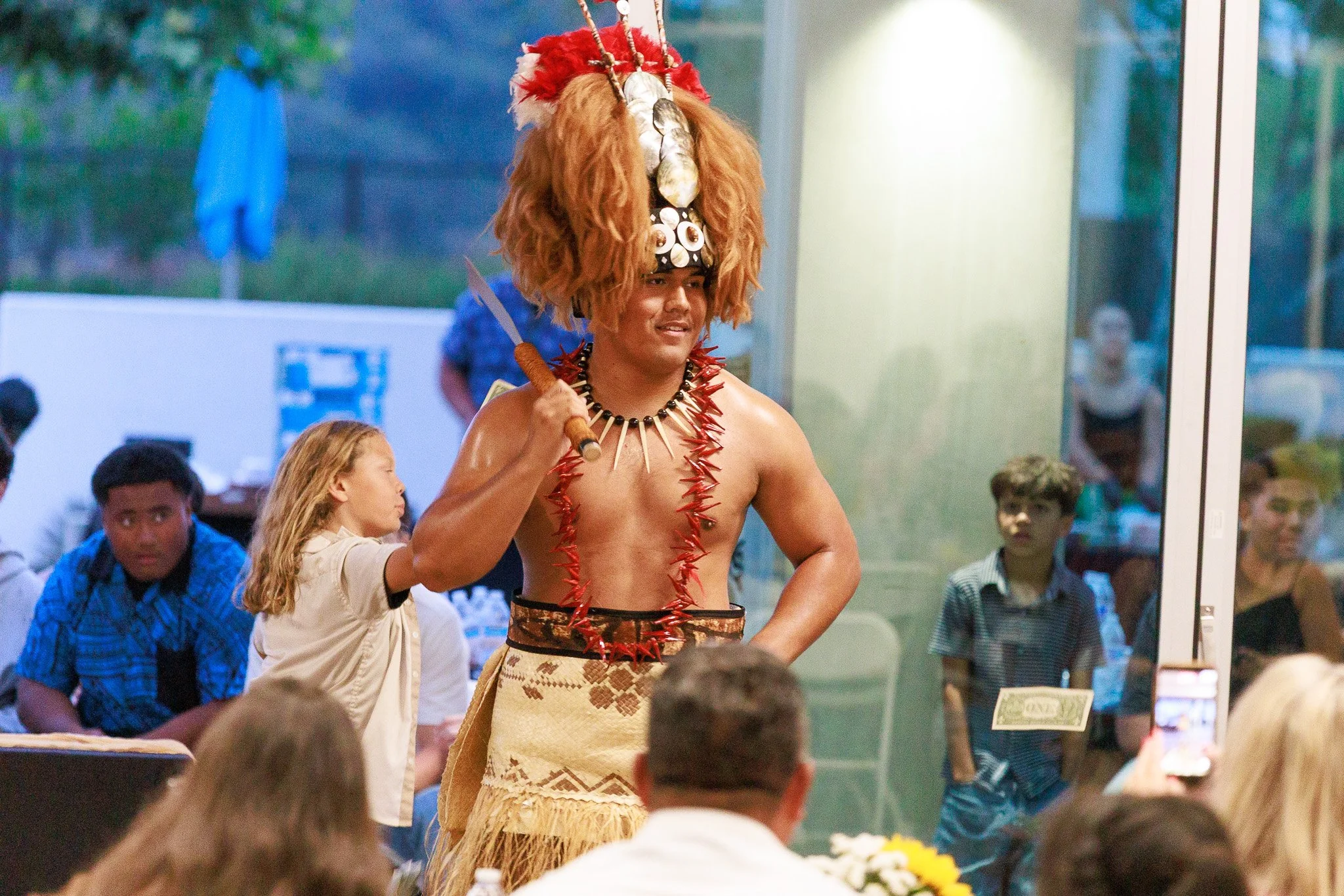 Cultural performer wearing a feathered headdress, shell jewelry, grass skirt, and holding a ceremonial knife before an audience