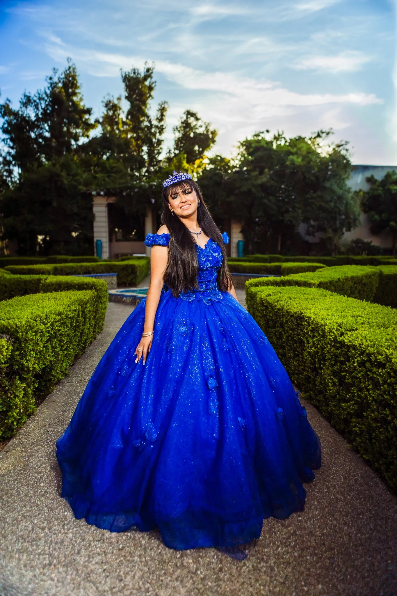 Young woman in a sparkling royal blue ball gown and tiara posing in a manicured garden.