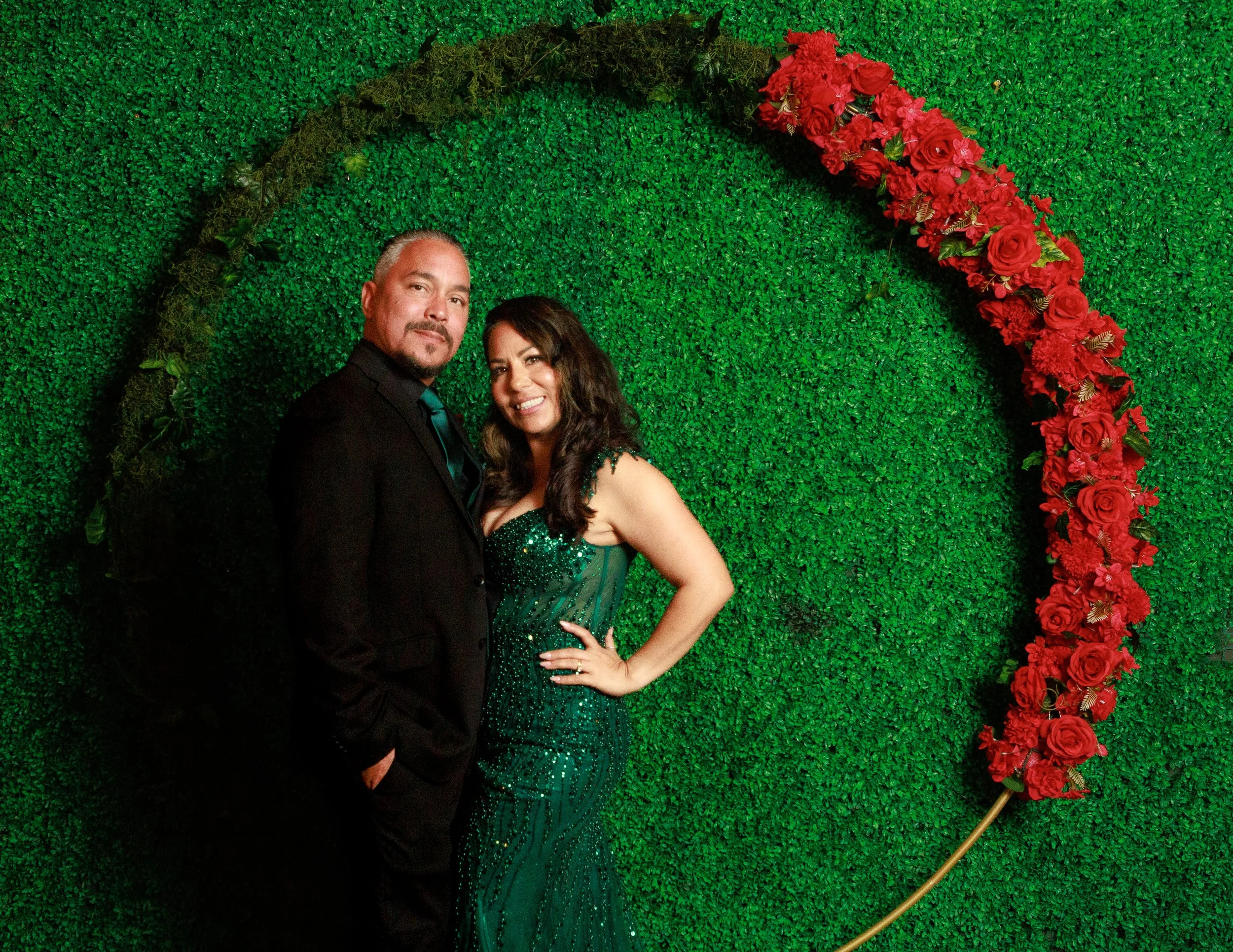 Couple posing in formal attire against a green foliage wall with red roses