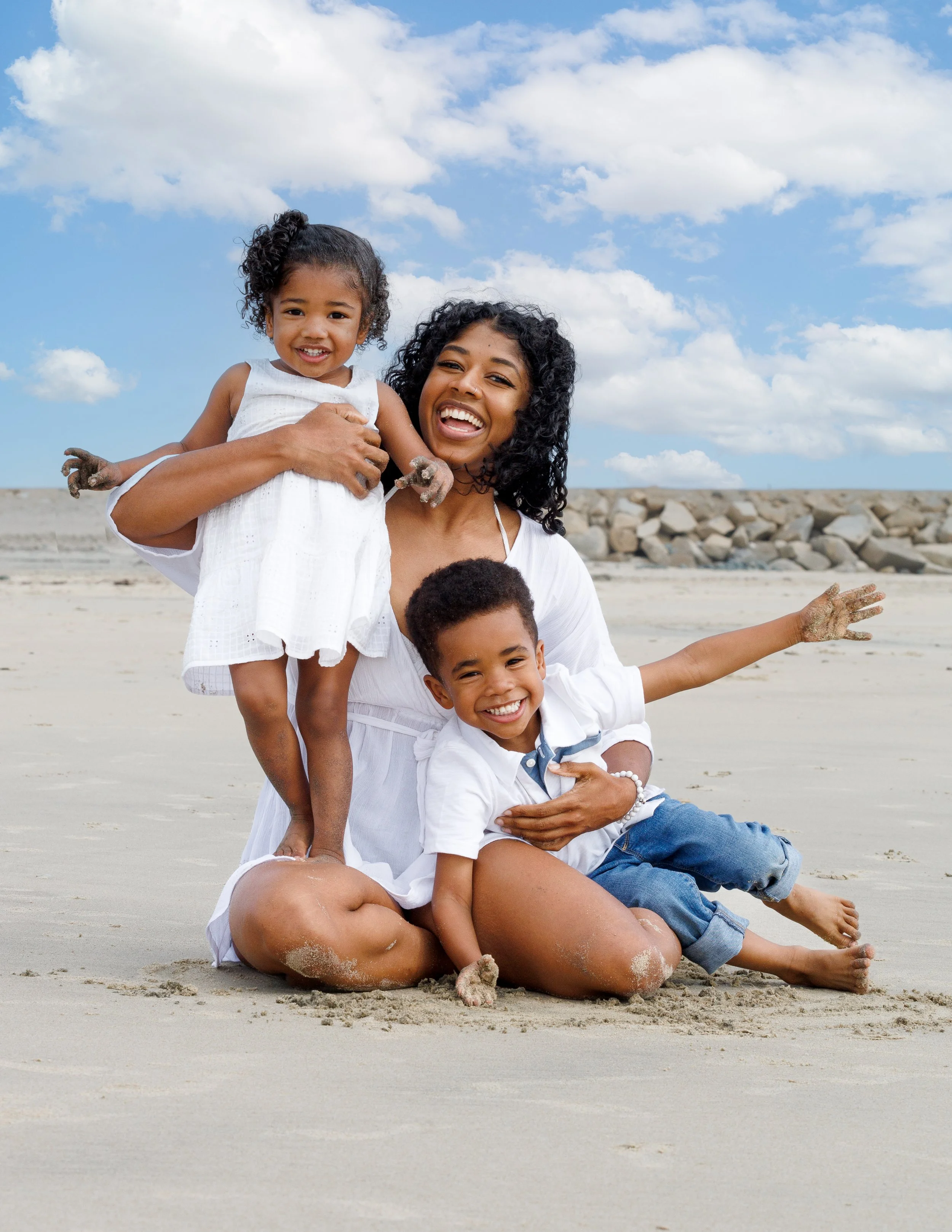 A group of happy diverse family members on the sandy beach smiling and playing together with a blue sky and scattered clouds in the background.