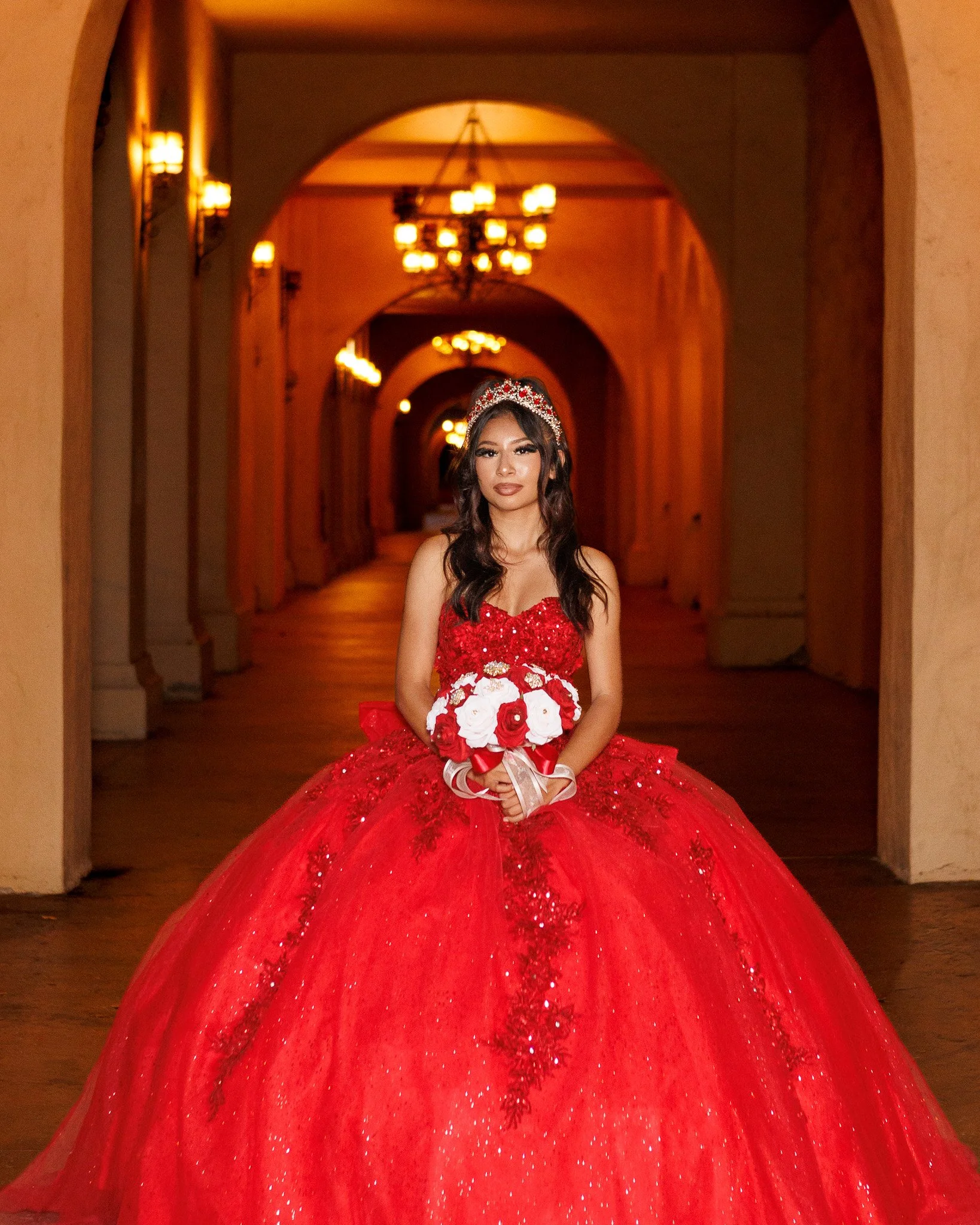 Young woman in a sparkling red quinceañera ball gown and tiara holding a bouquet in an ornate, warmly lit hallway