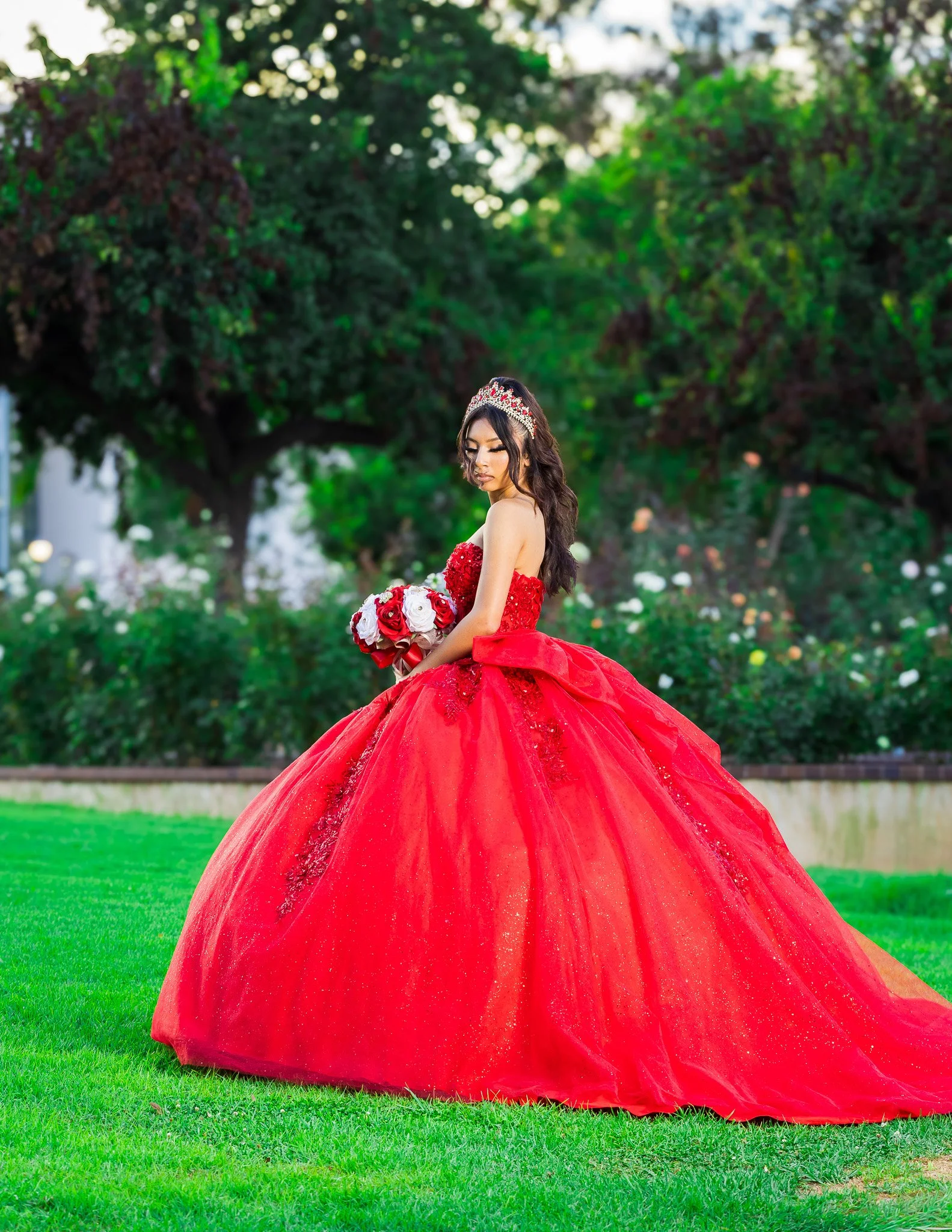 Young woman in a sparkling red ball gown and tiara holding a red and white rose bouquet in a garden