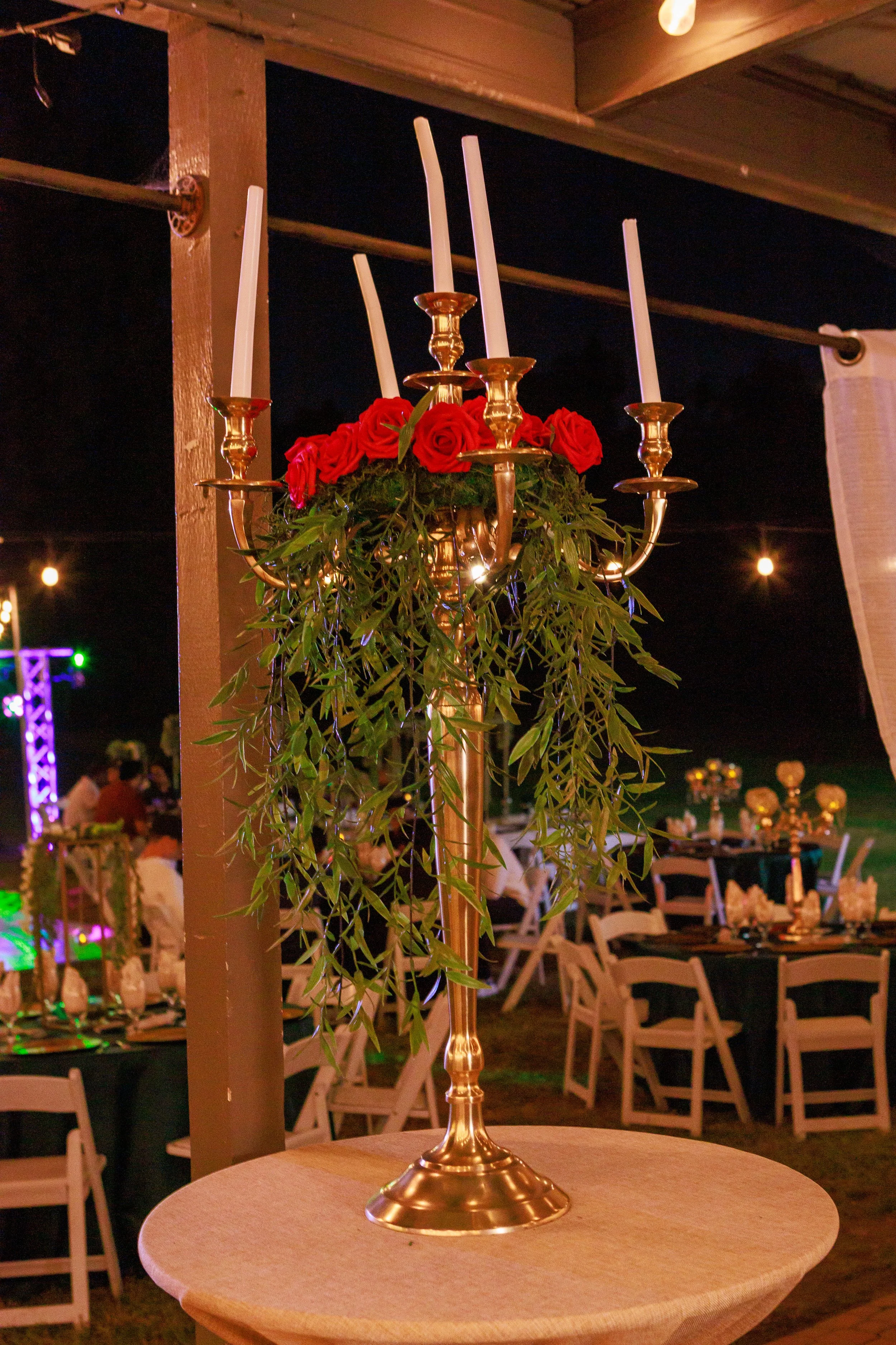Elegant outdoor wedding table centerpiece with gold candelabra, white taper candles, red roses, and cascading greenery.
