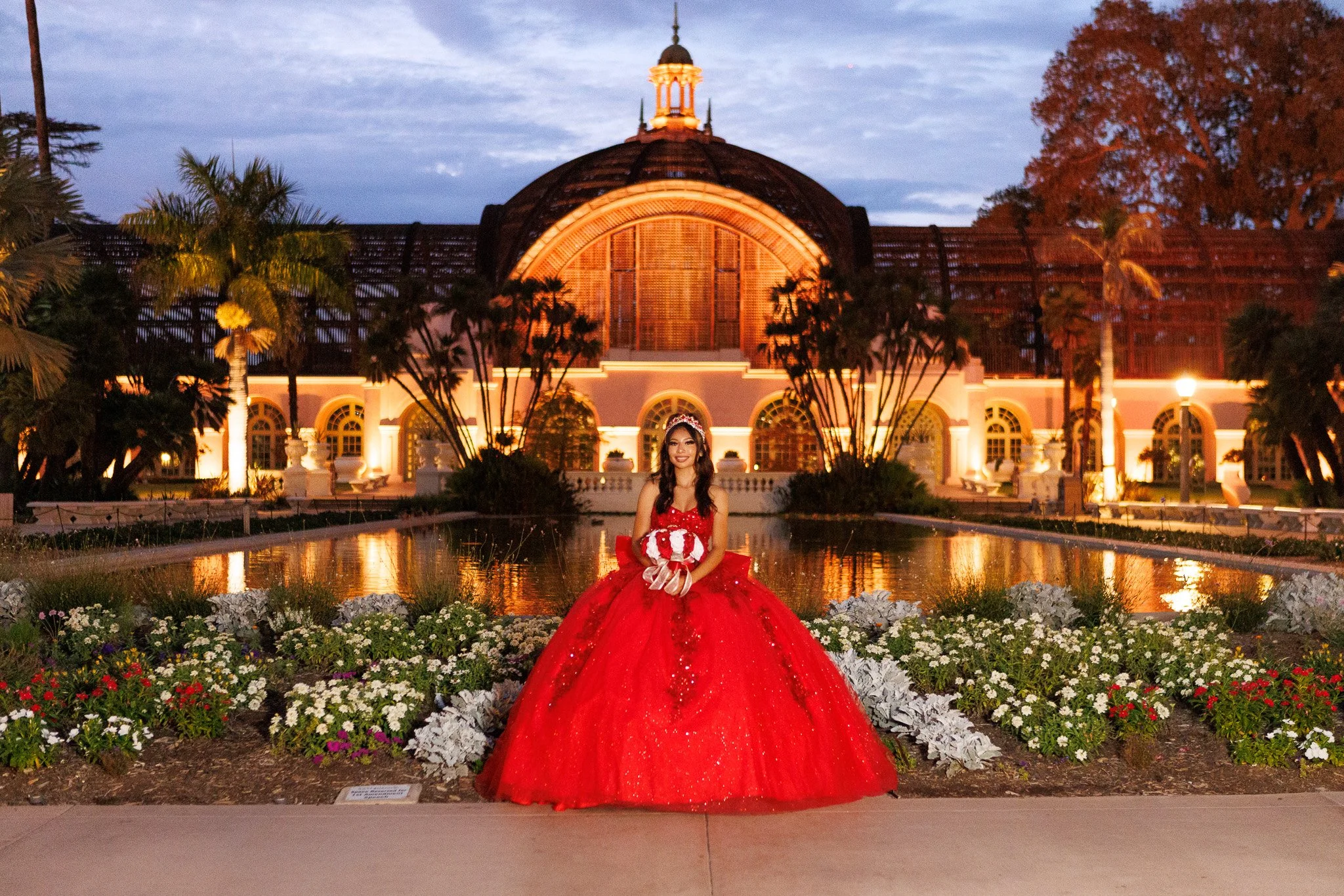 Woman in a sparkling red ball gown holding a bouquet in front of an illuminated conservatory and reflecting pool at dusk