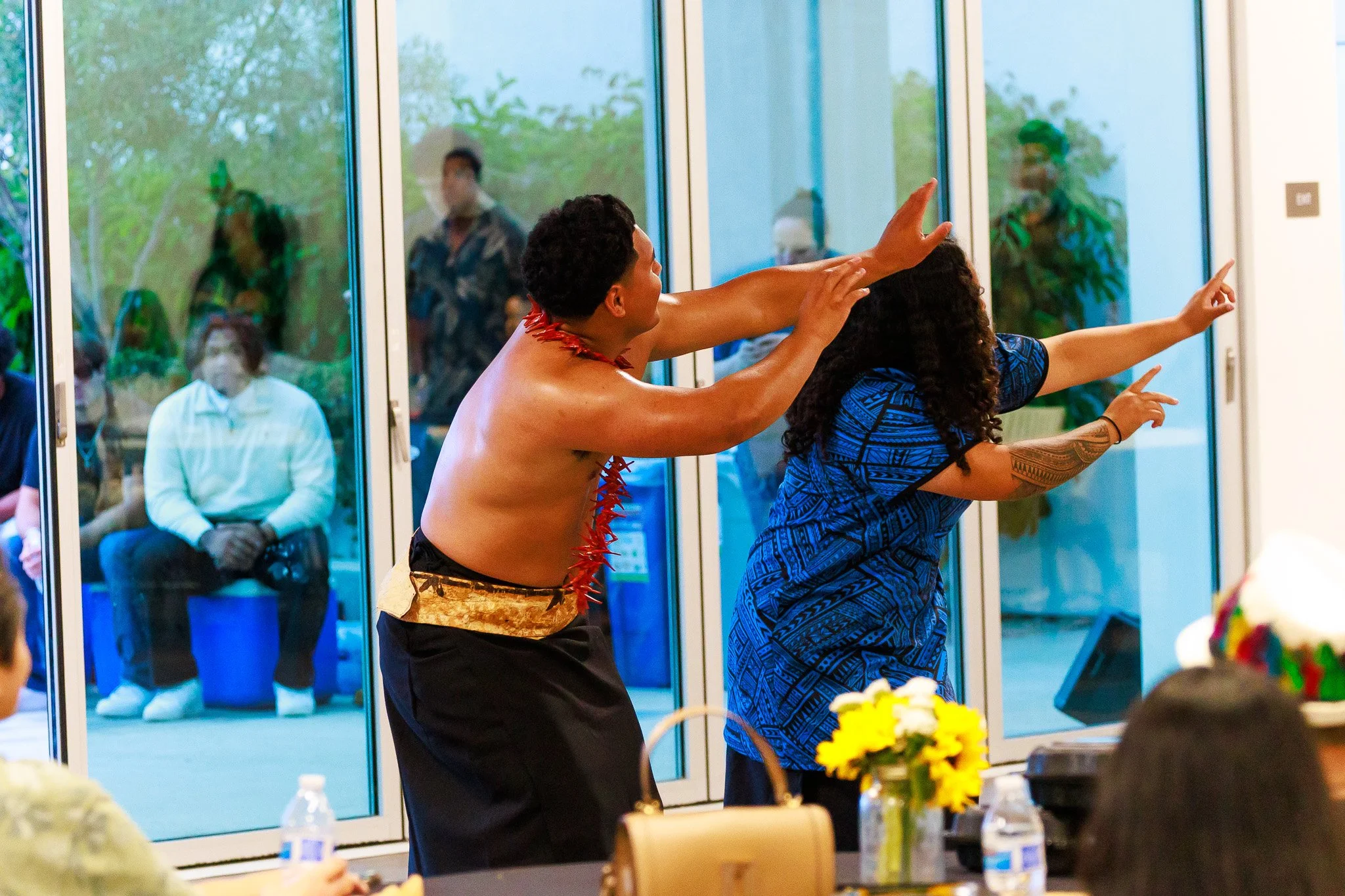 Two performers in traditional attire dance with outstretched arms at an indoor cultural event.