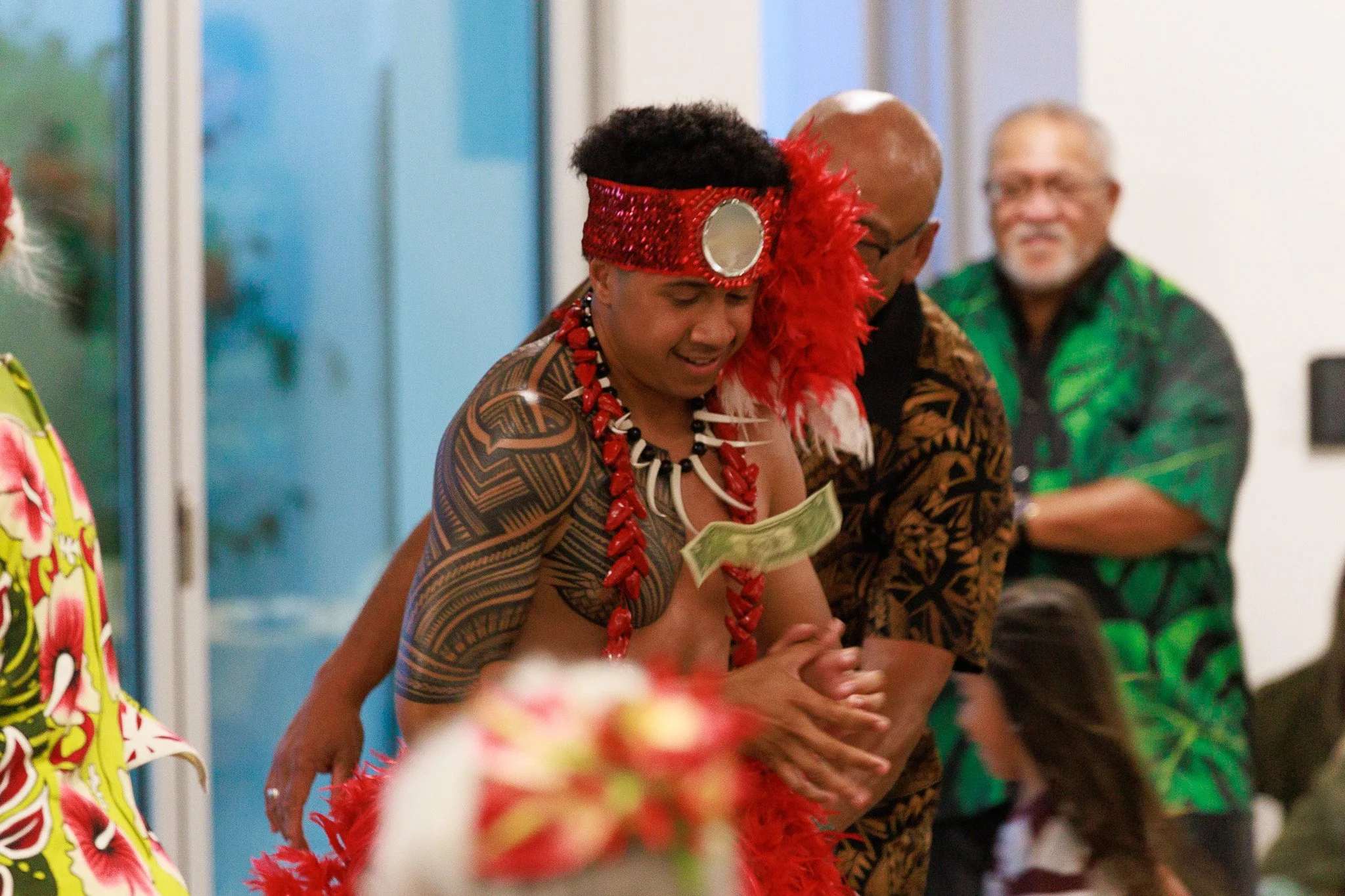 Person in traditional Polynesian attire with red feathered headpiece and lei, surrounded by people at a cultural celebration
