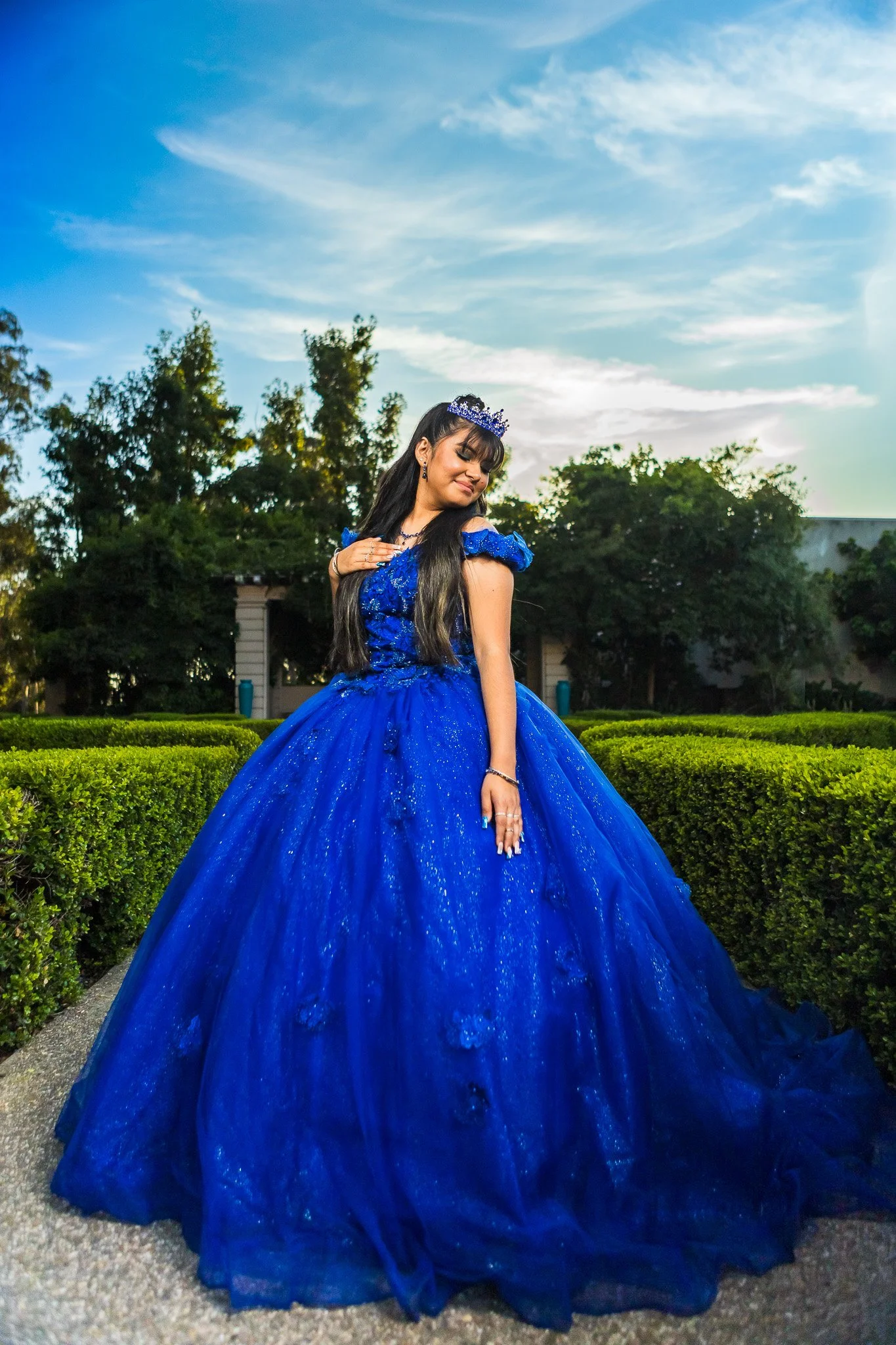 Young woman wearing a sparkling royal blue ball gown and tiara outdoors in a landscaped garden