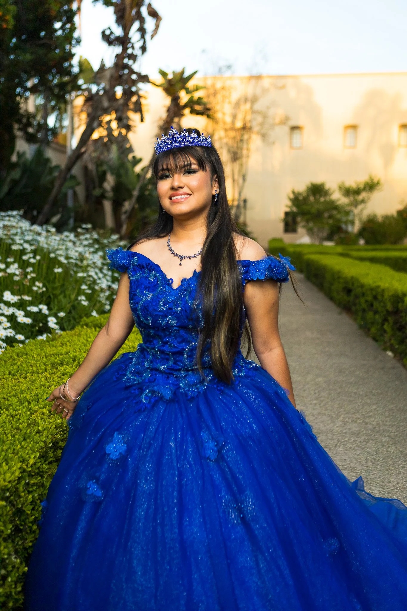 Smiling woman in a jeweled blue tiara and embellished royal blue ball gown posing in a garden