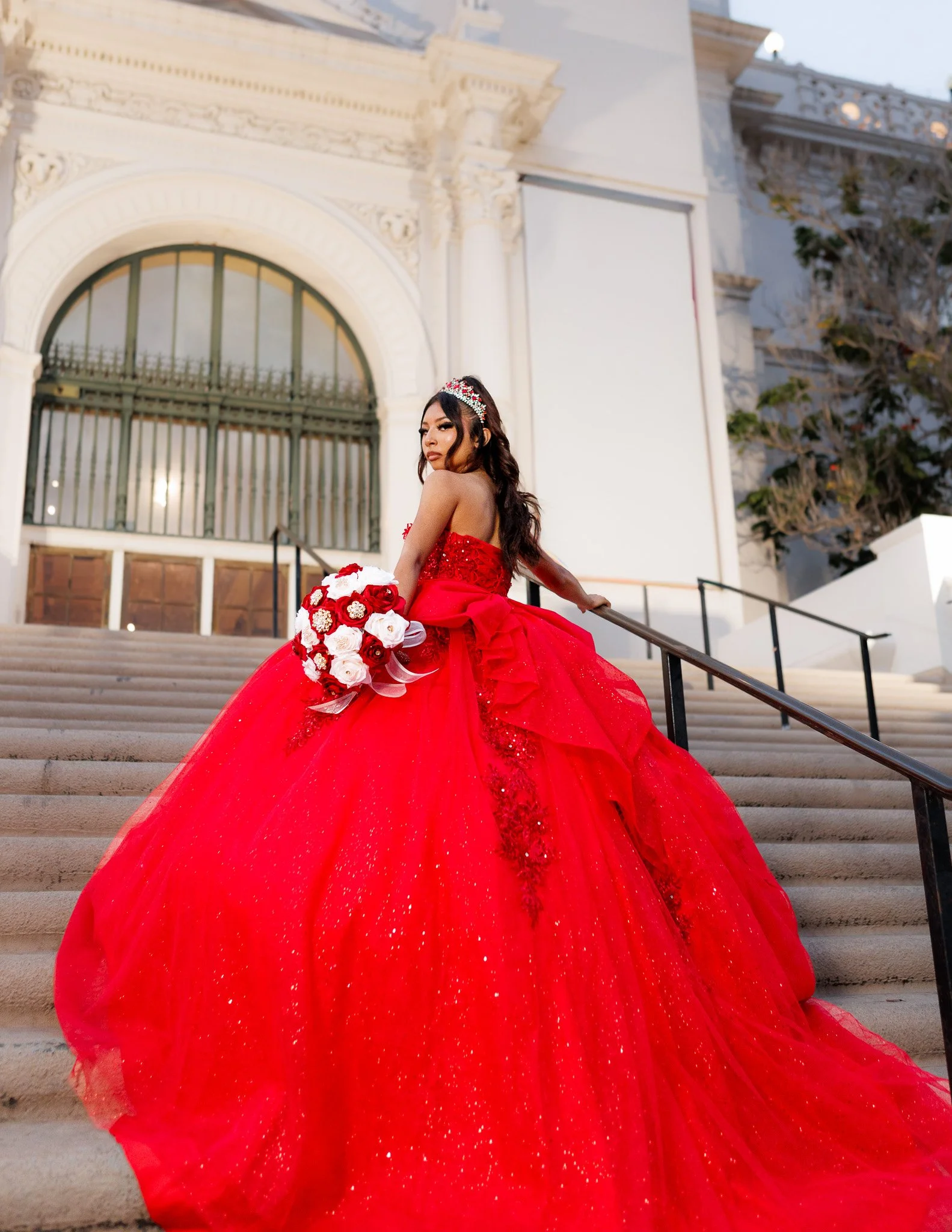 Quinceañera in a sparkling red ball gown holding a red and white bouquet on grand courthouse steps