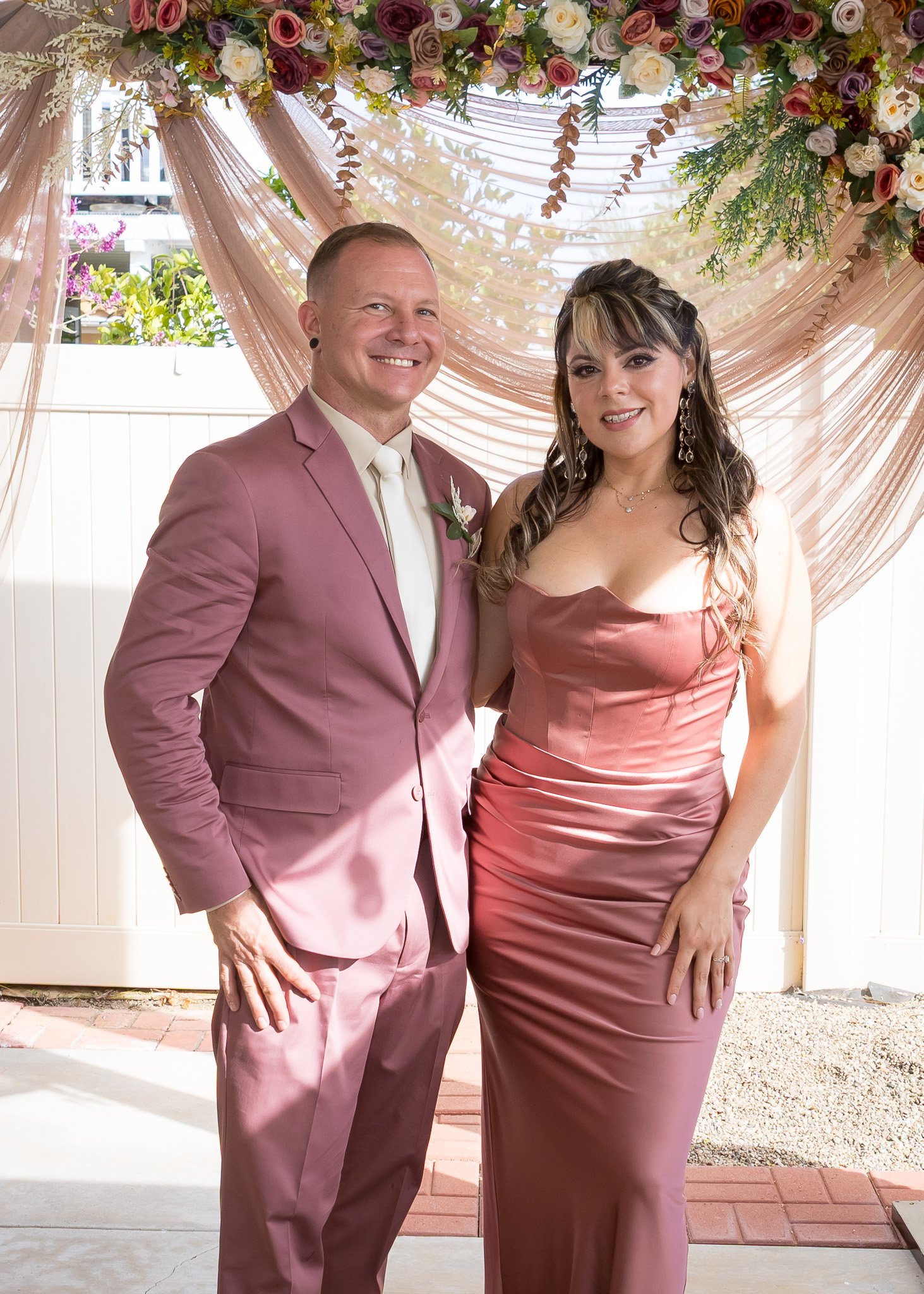 A man and woman dressed in pink formal attire, standing closely together under a decorative floral backdrop at a wedding or special event, smiling at the camera.