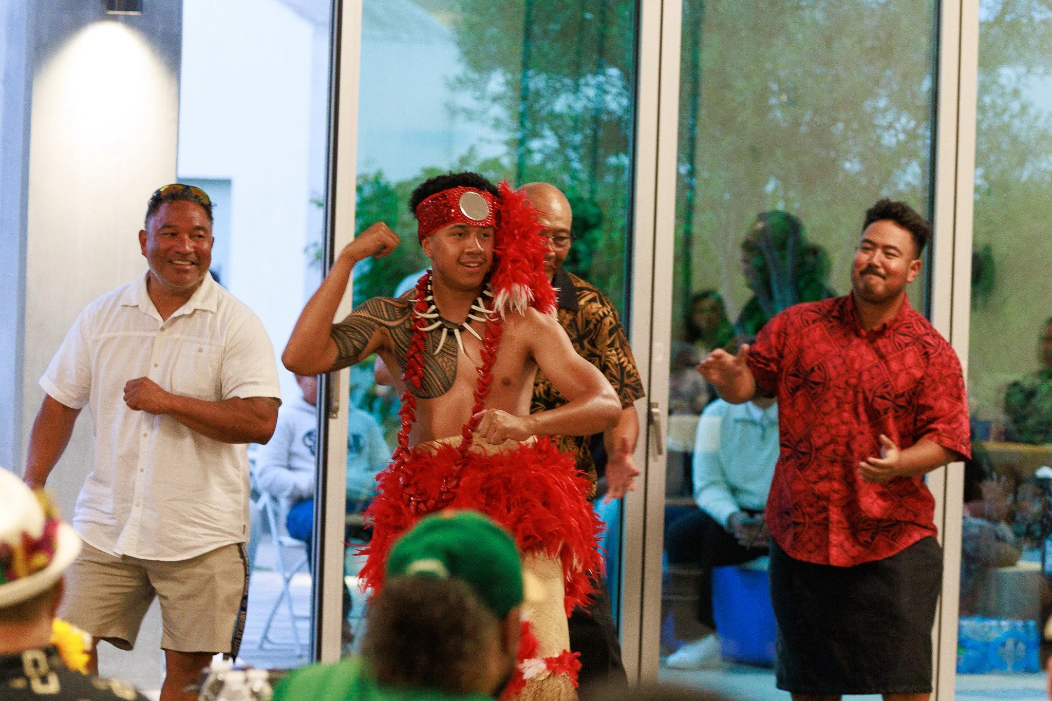 People performing a lively Polynesian dance indoors, featuring a dancer in a red feathered costume and traditional headpiece.