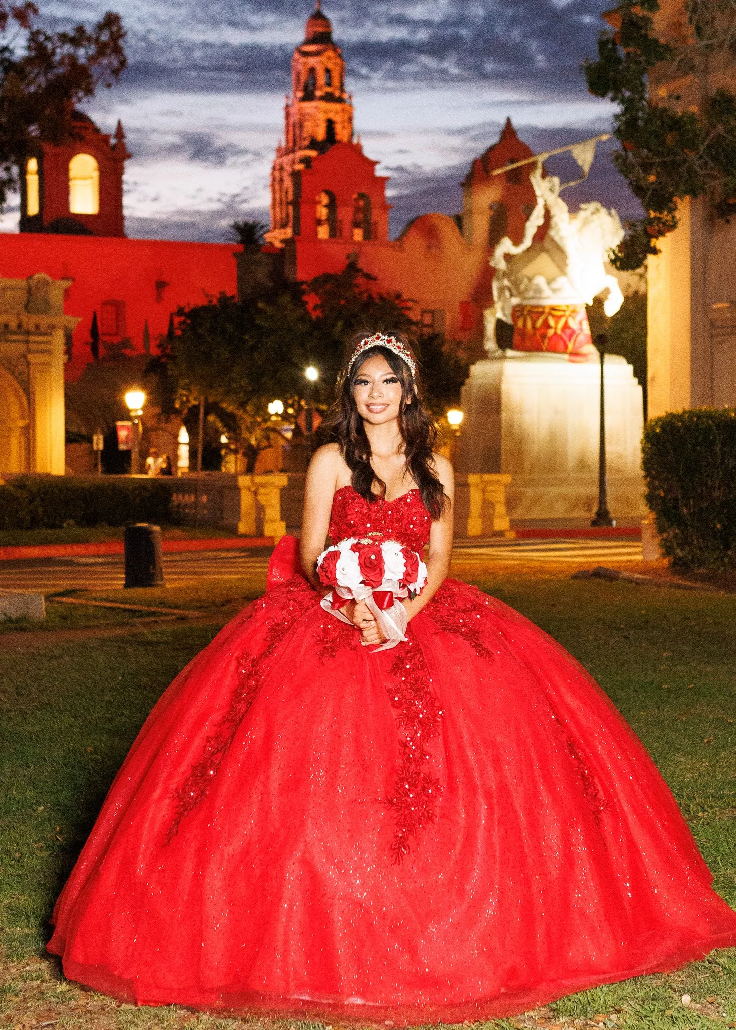 Young woman in a red quinceañera ball gown holding a bouquet in front of illuminated historic buildings at dusk
